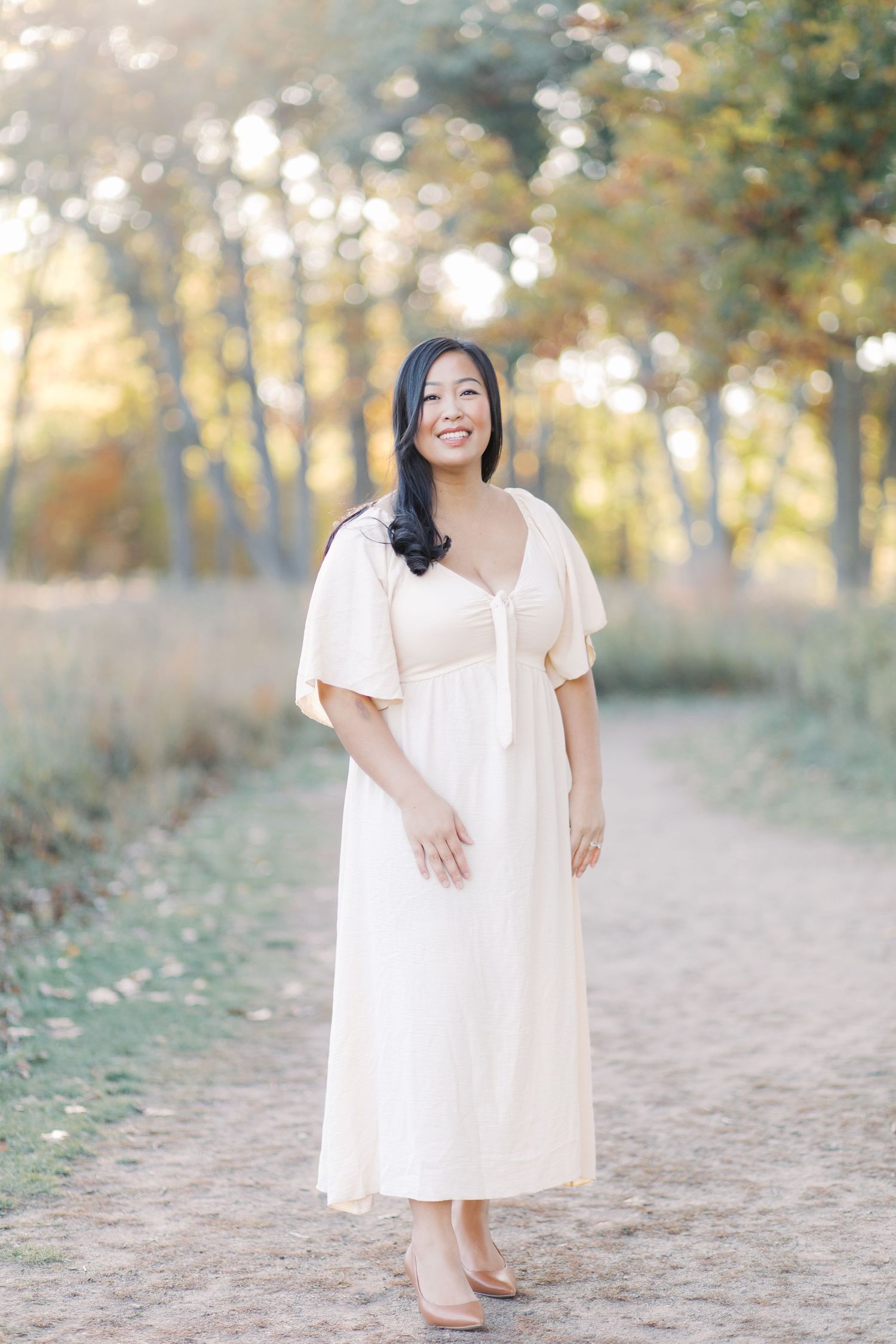 A woman in a flowing white dress stands on a dirt path surrounded by autumn trees at golden hour.