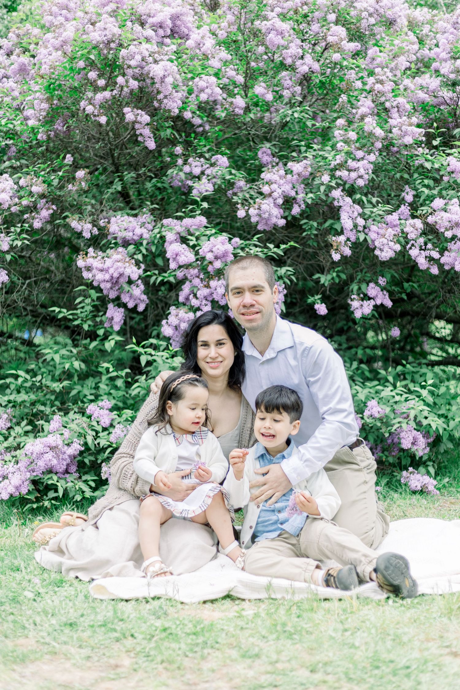Family enjoying a spring day together on a blanket surrounded by blooming pink lilac bushes in a garden setting.