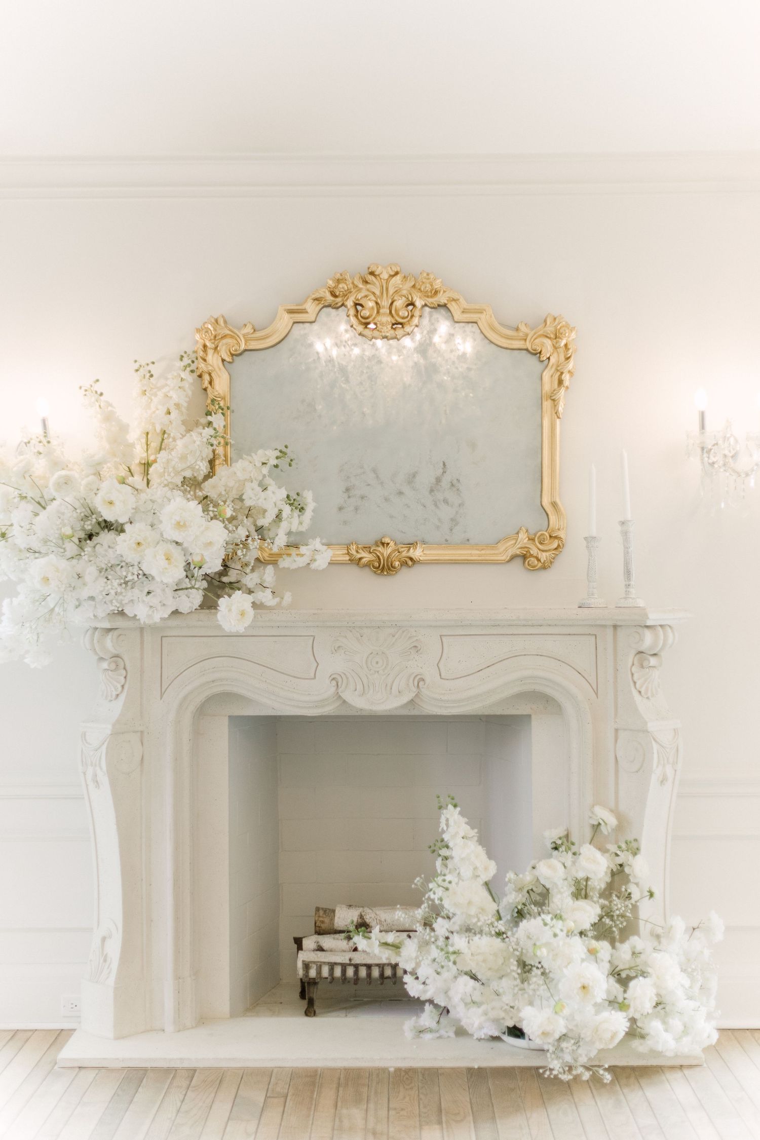 Romantic white interior featuring decorative gold-framed mirror and delicate white flower arrangements above a fireplace.