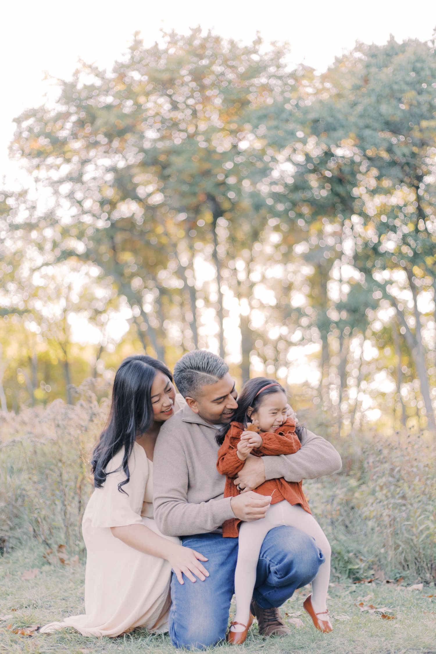 A family shares tender moments together in a sunlit outdoor setting with autumn trees in the background.