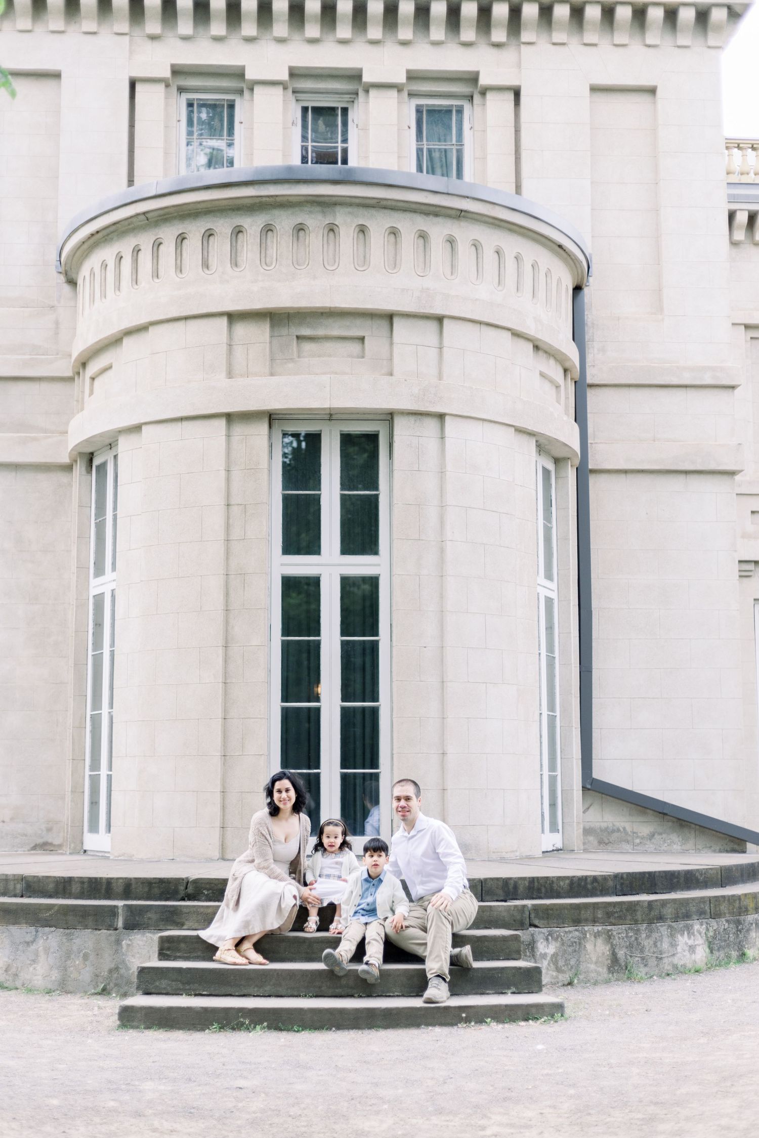 Small family sitting together on stone steps in front of a grand white building with curved architectural features.