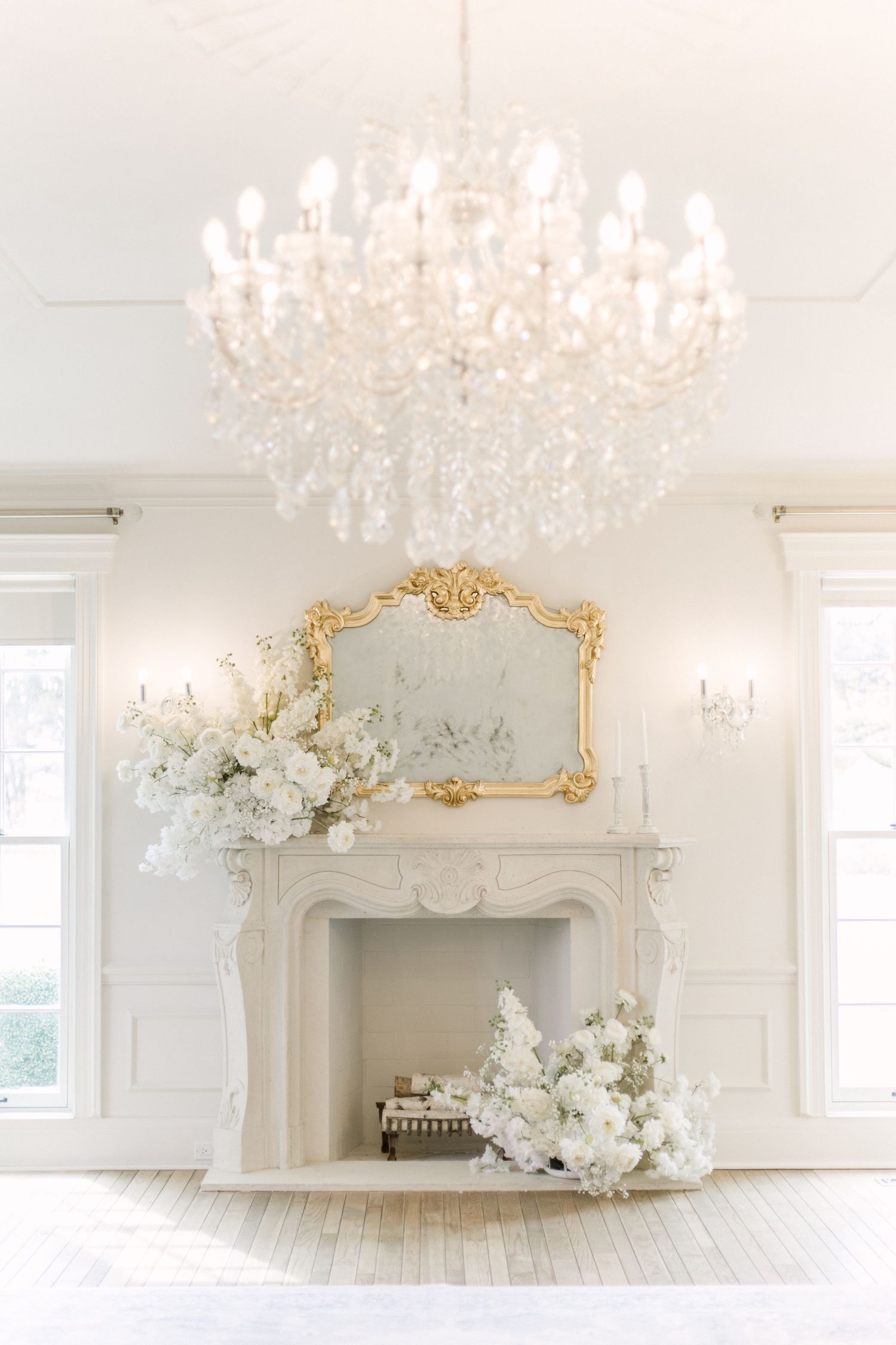 Elegant white room with crystal chandelier, ornate gold mirror above fireplace, and white floral arrangements.