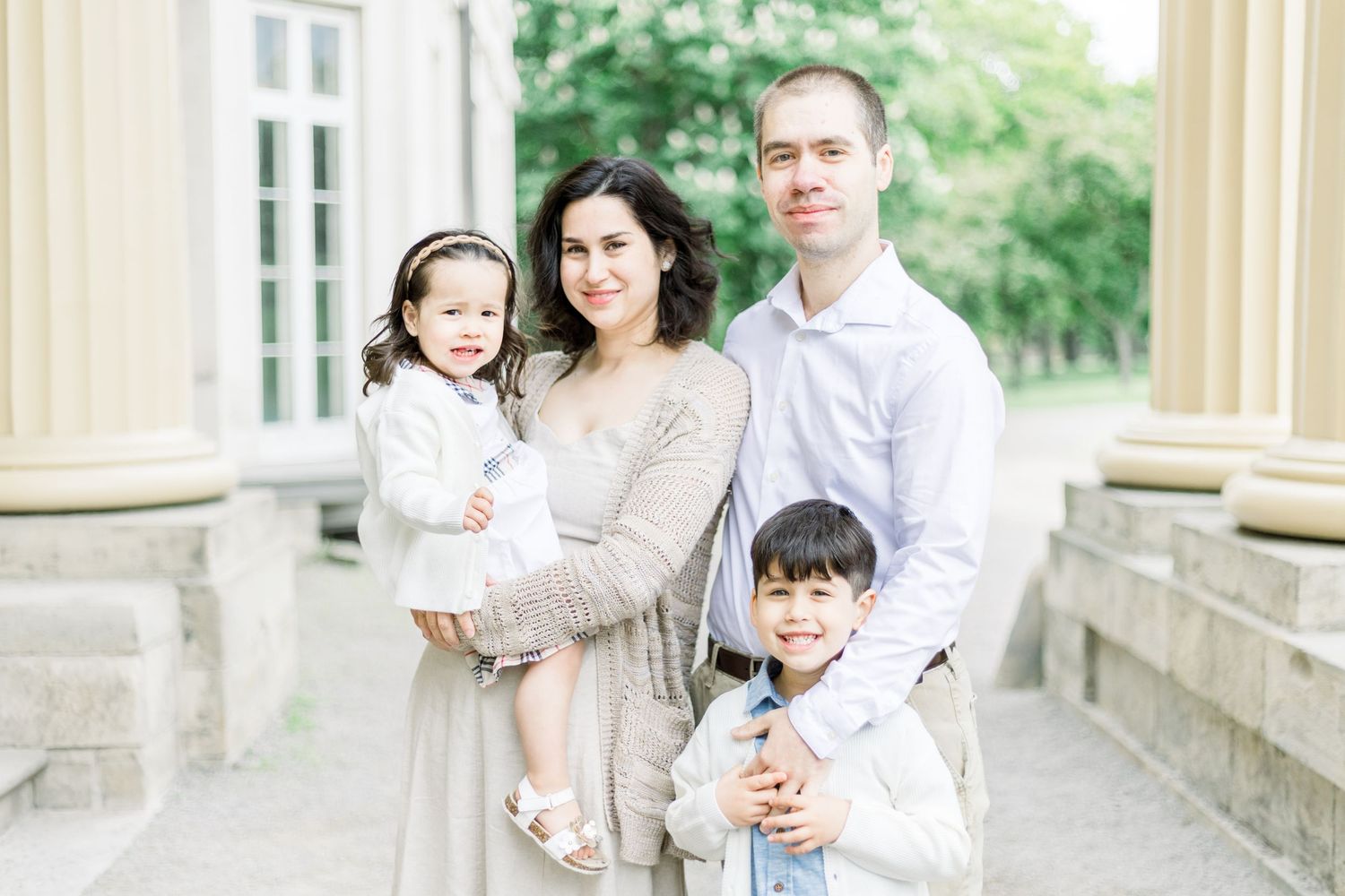 A family of four wearing neutral colored clothing poses together between stone columns outdoors.