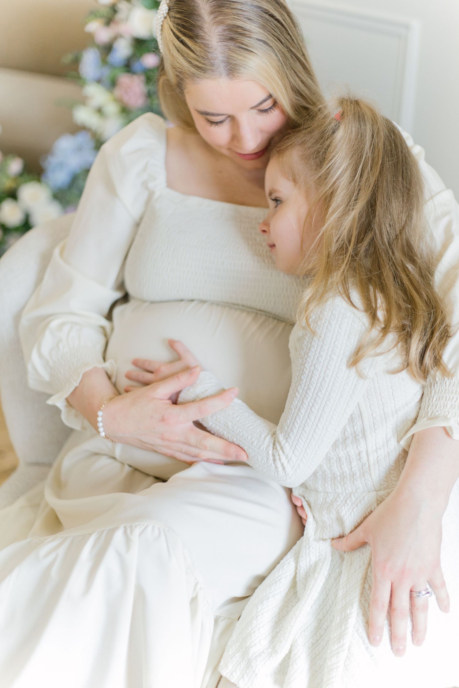 A tender pregnancy photoshoot series showing a woman in white sitting indoors with soft lighting and floral decor.