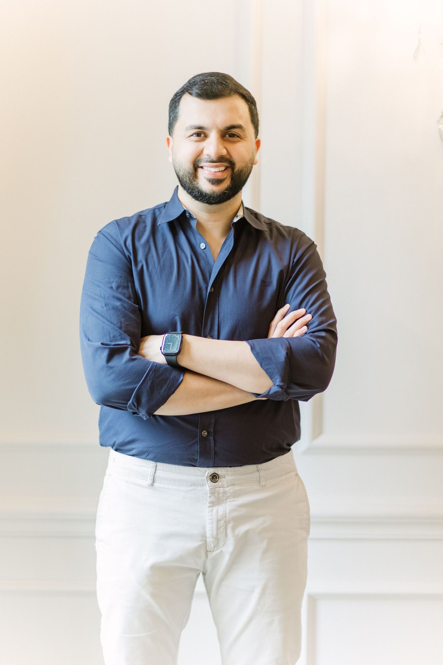 Professional wearing a navy blue button-down shirt and white pants stands with arms crossed in a bright studio setting.