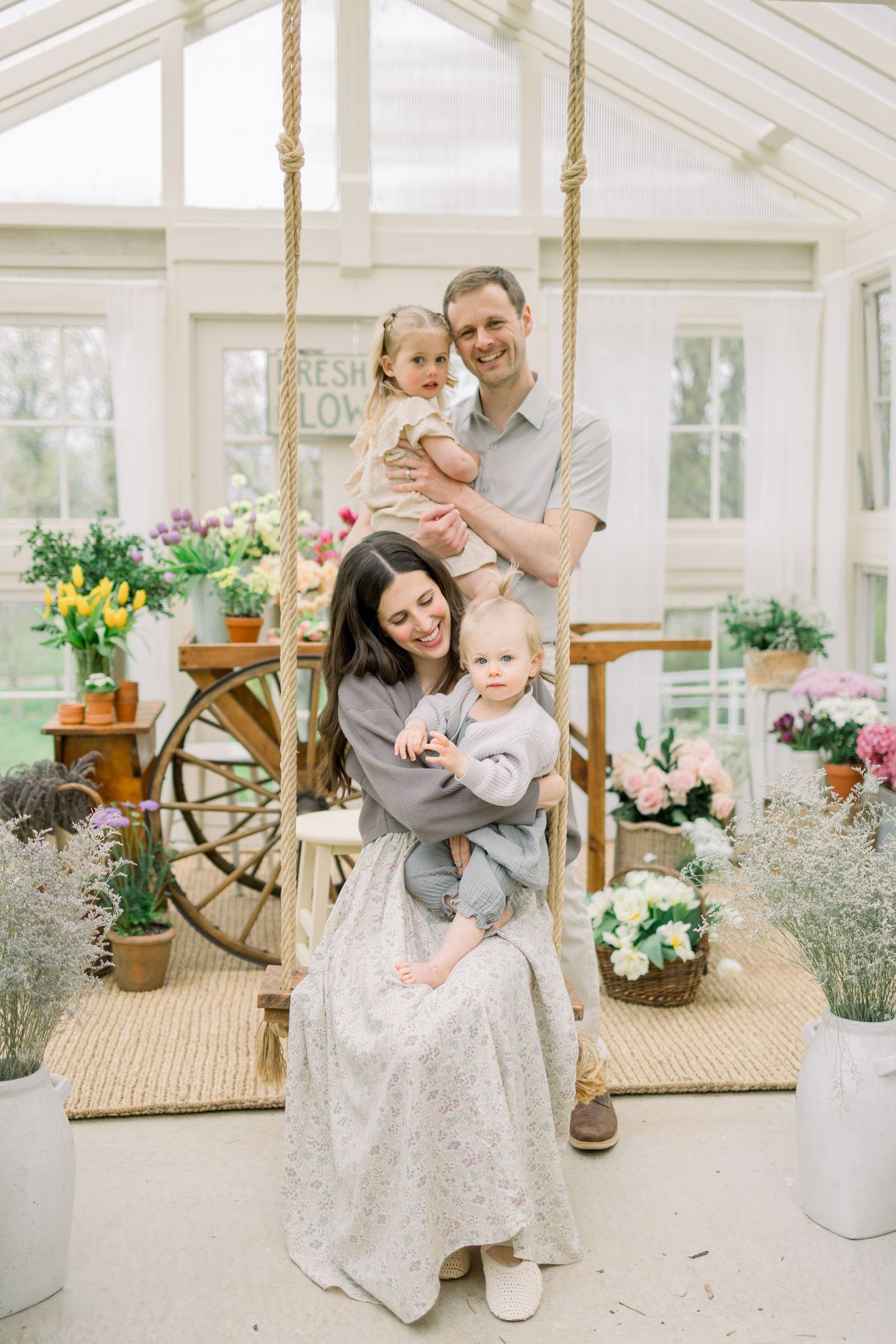 Family shares tender moments together in a bright sunlit conservatory filled with potted plants and flowers.