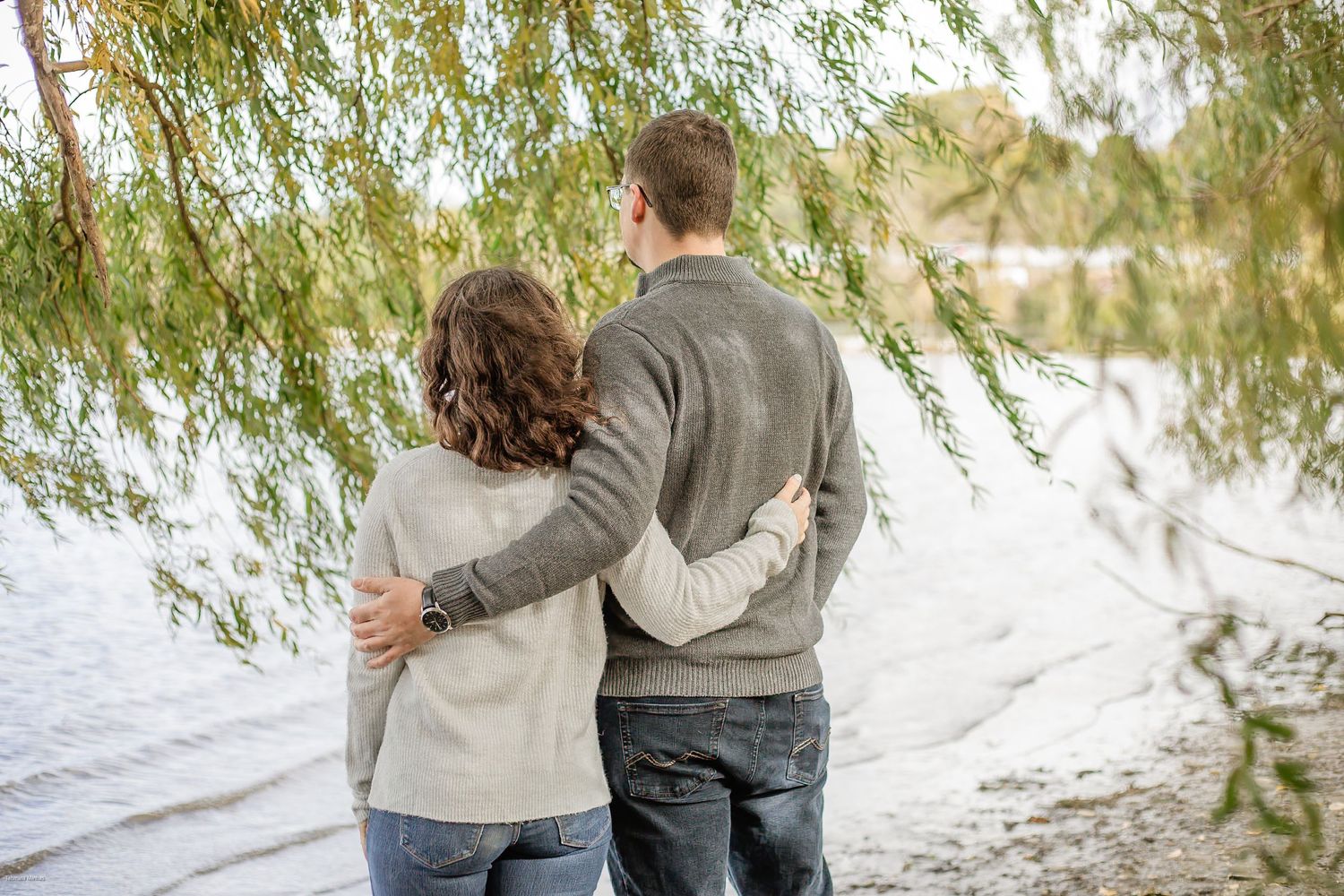 Couple stands close together looking at lake view through willow tree branches on peaceful autumn afternoon.