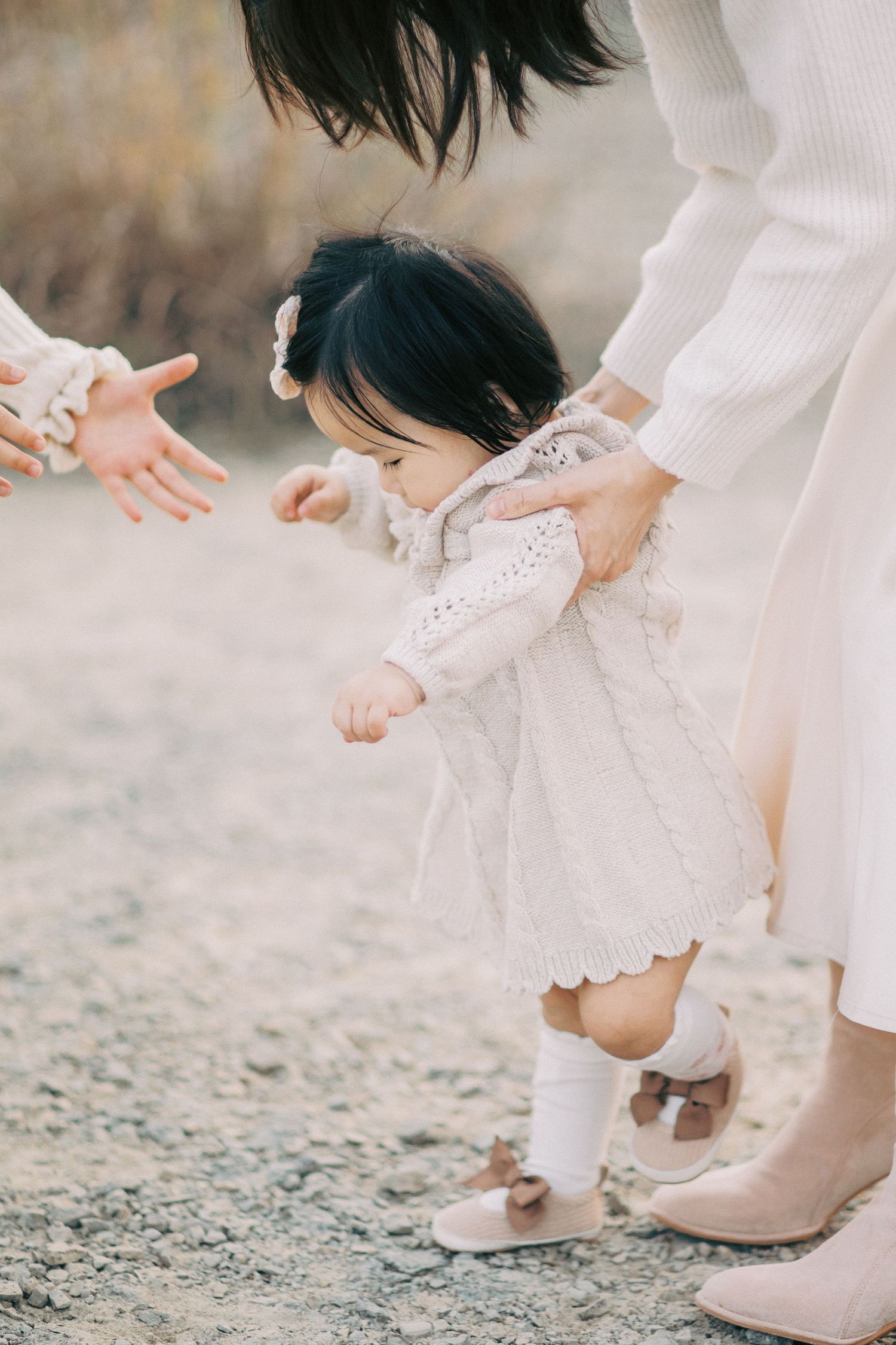 Close-up shots of a child's feet in white shoes and dress taking first steps on gravel with adult hands guiding.