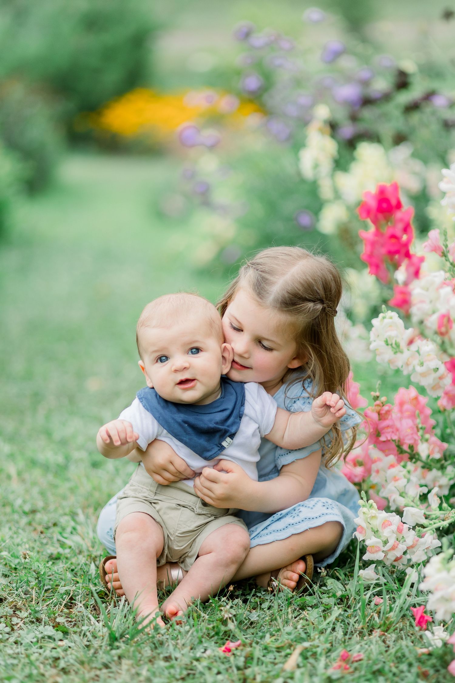 Two children sit together among colorful spring flowers in a garden setting, sharing a tender moment.