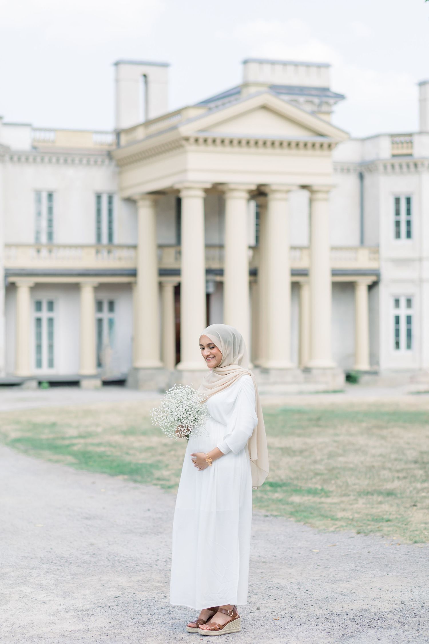 A person in a flowing white dress and hijab holds baby's breath flowers while standing in front of a neoclassical building.