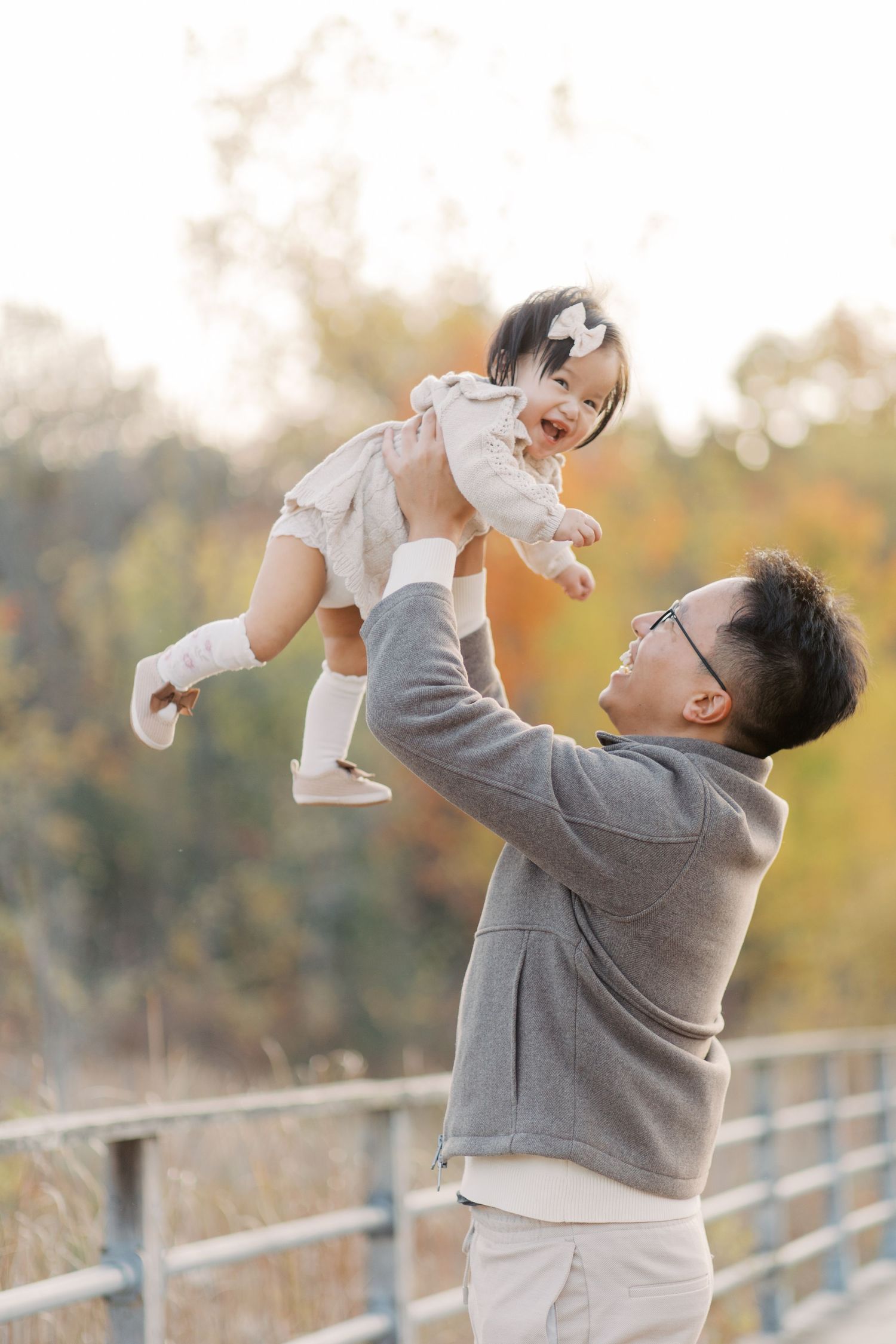 A heartwarming autumn scene shows a parent lifting a baby wearing white into the air against fall foliage.