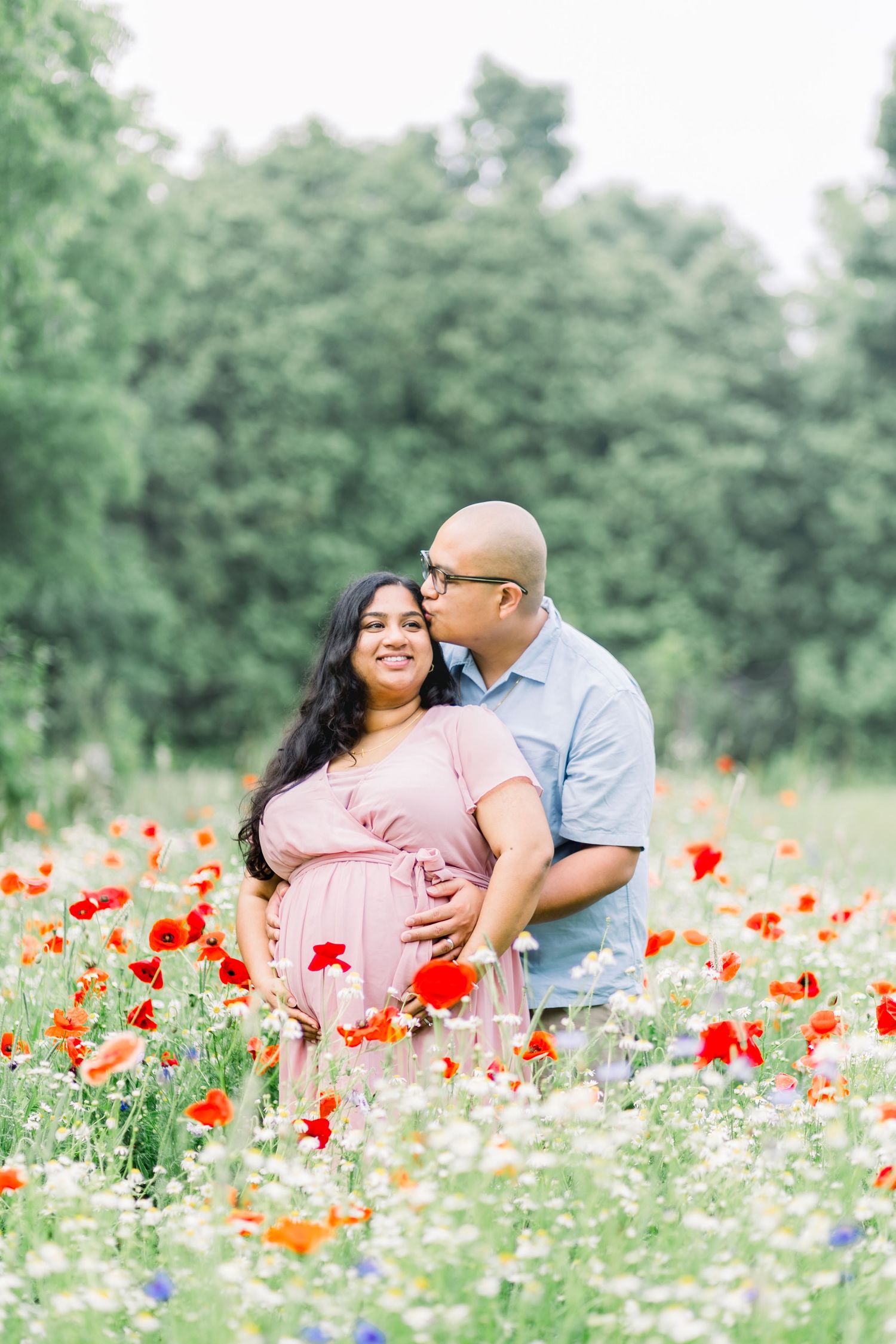 A couple shares tender moments while laying in a vibrant field of red poppies and white wildflowers on a summer day.