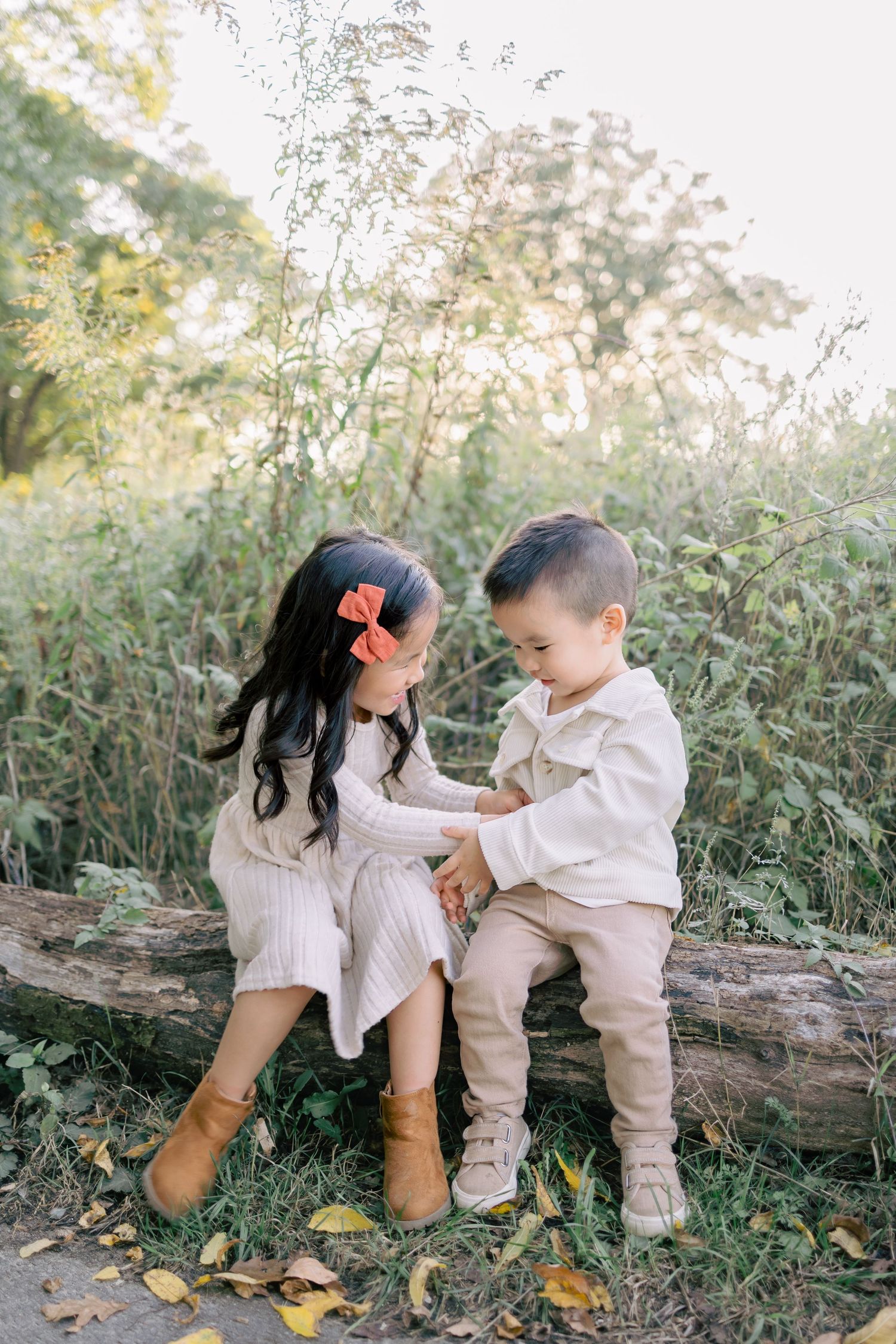 Two children sit together on a fallen log surrounded by autumn foliage and natural greenery.