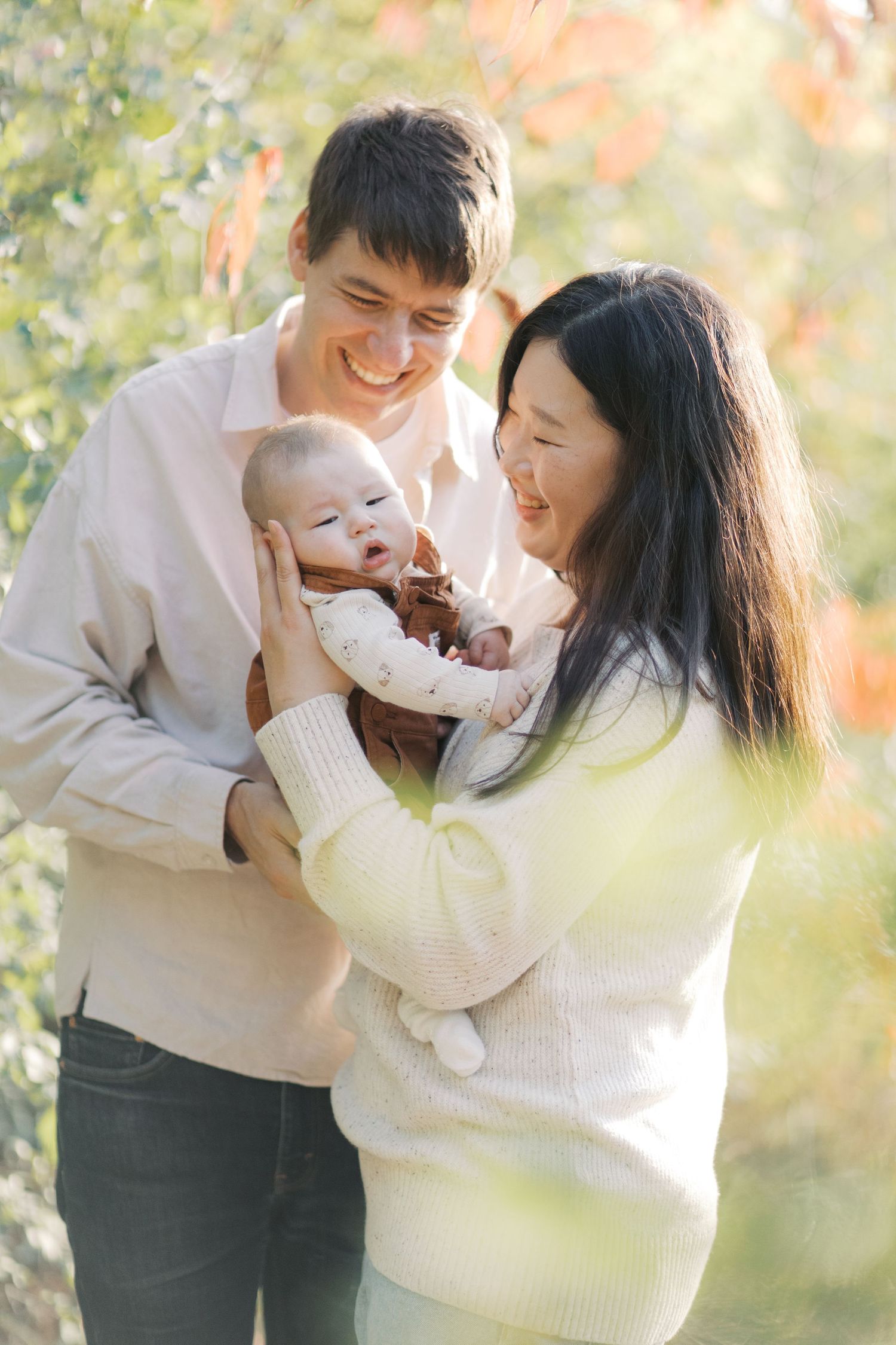 A couple in white sweaters share joyful moments with their baby outdoors on an autumn day.