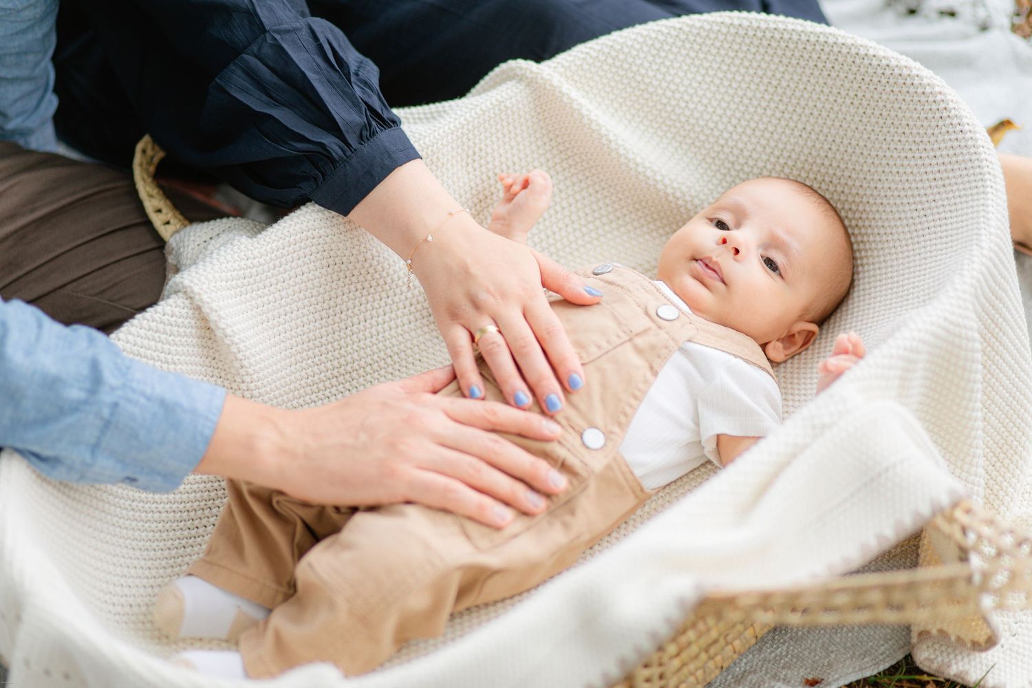 A small baby in white clothes lies on a cream-colored blanket while hands gently rest on their tummy.