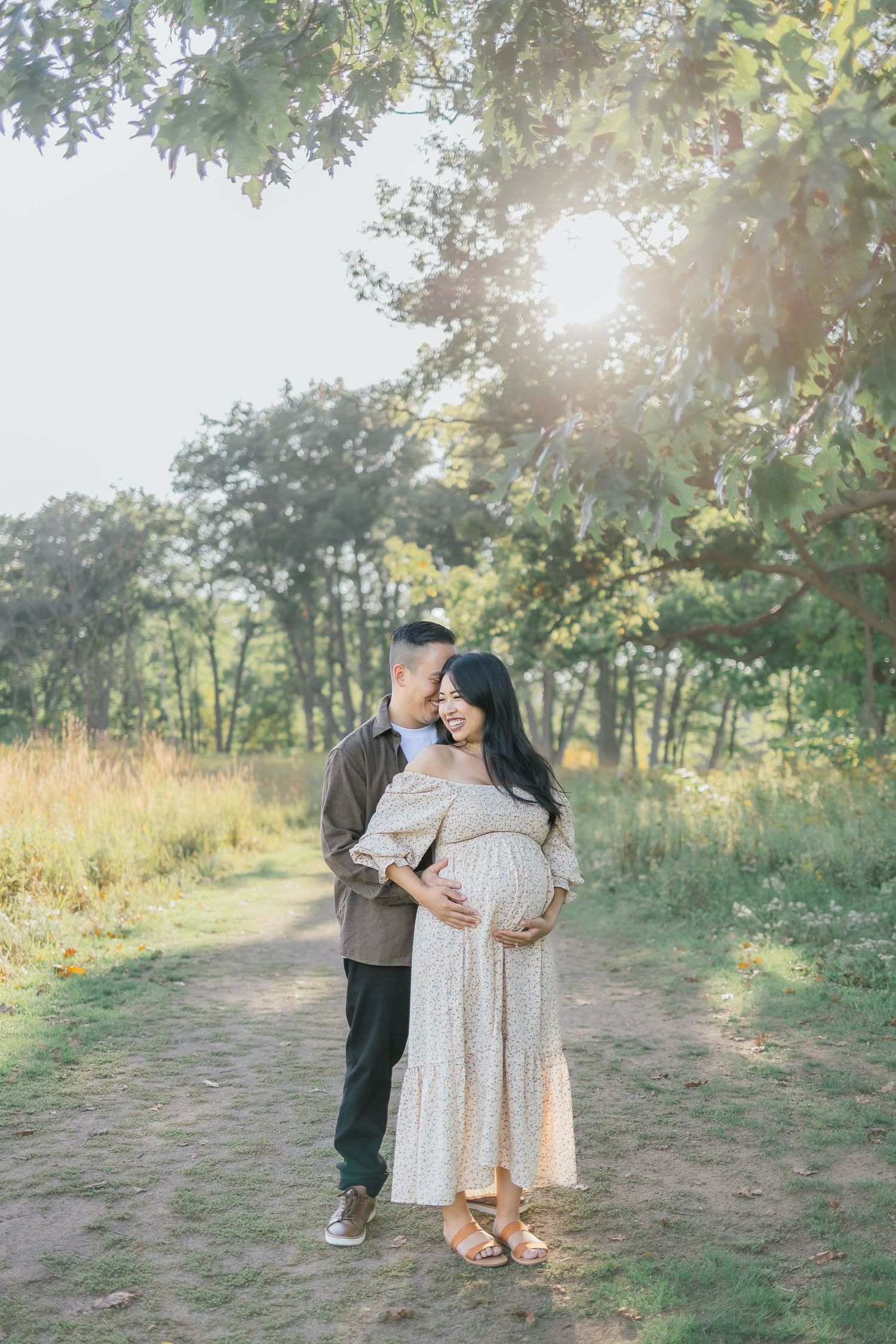 A couple shares romantic moments on a sunlit nature trail surrounded by trees and tall grass.