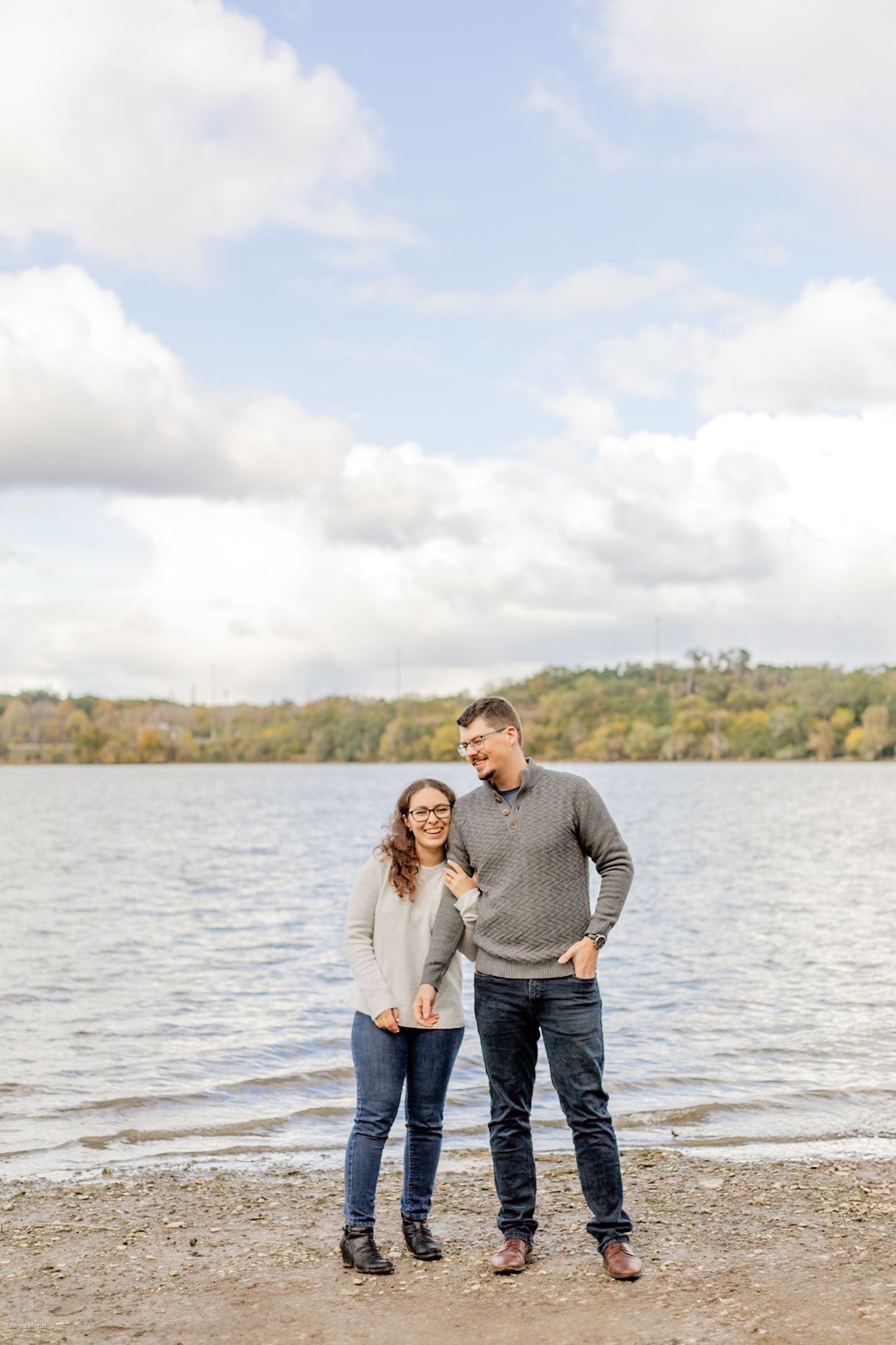 A couple stands together on a beach shoreline by a lake on an autumn day with cloudy skies overhead.