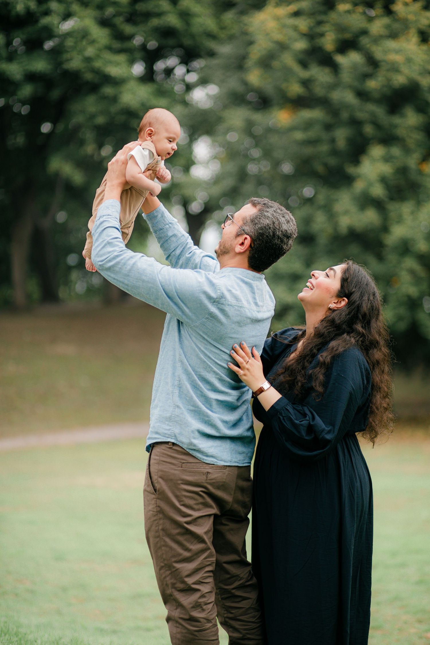 A couple joyfully plays with their baby on a grassy lawn surrounded by trees on a sunny day.
