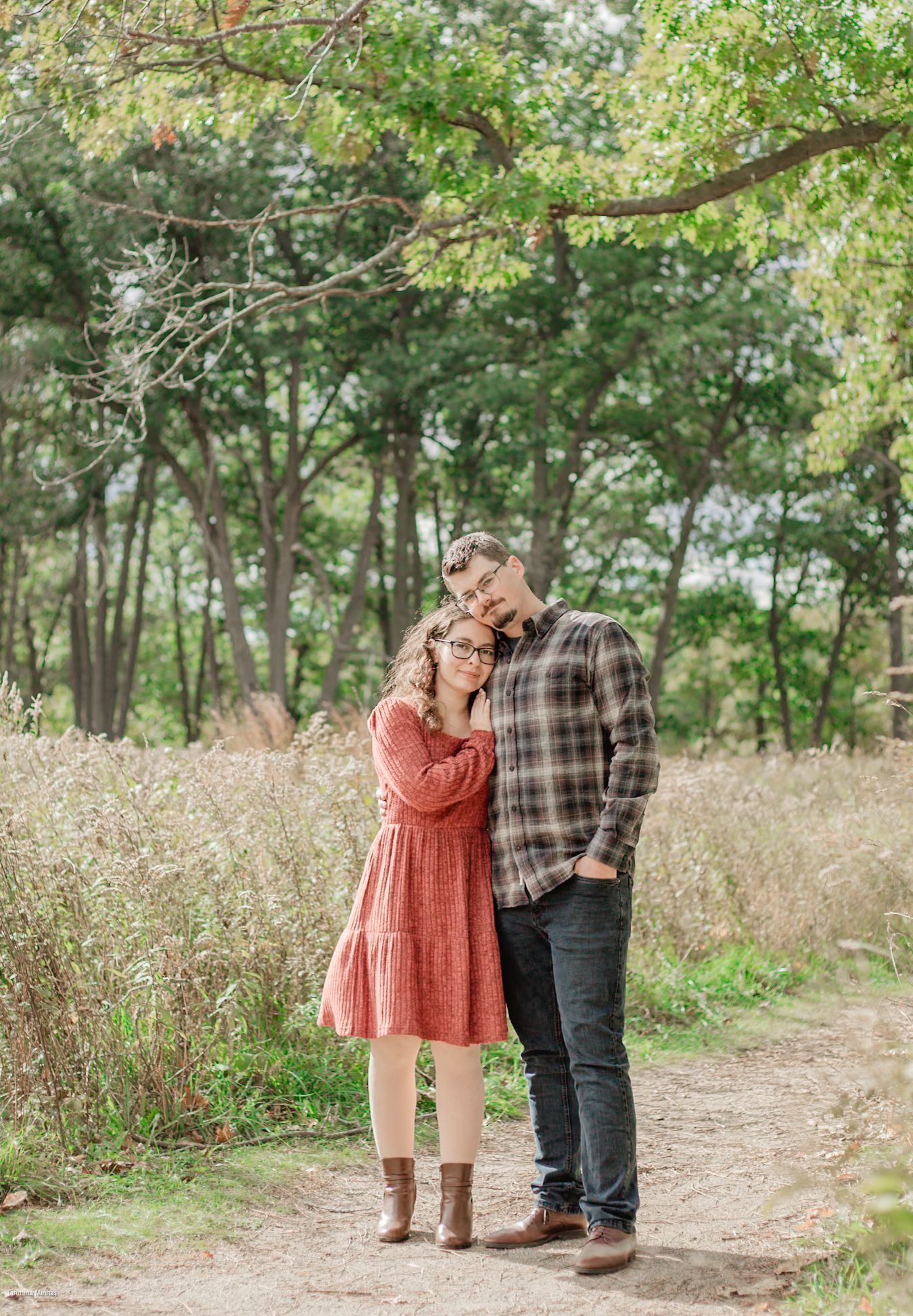 A couple shares a romantic moment while standing on a dirt path surrounded by tall grass and trees in autumn.