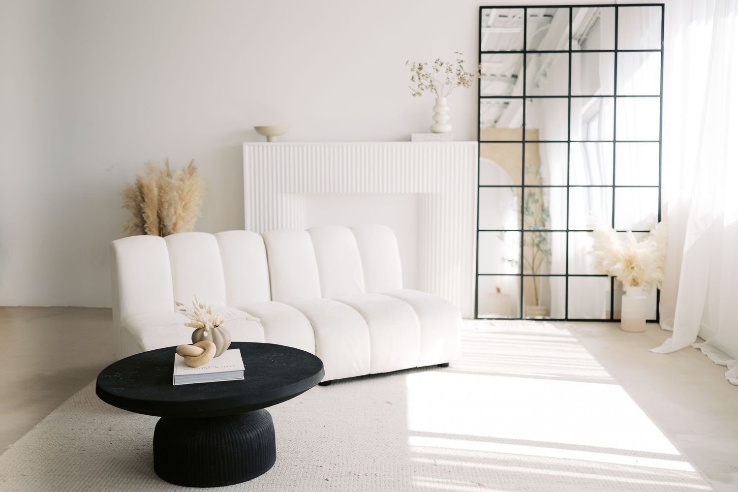 Modern minimalist white living room with black coffee table, window grid mirror and natural light streaming in.