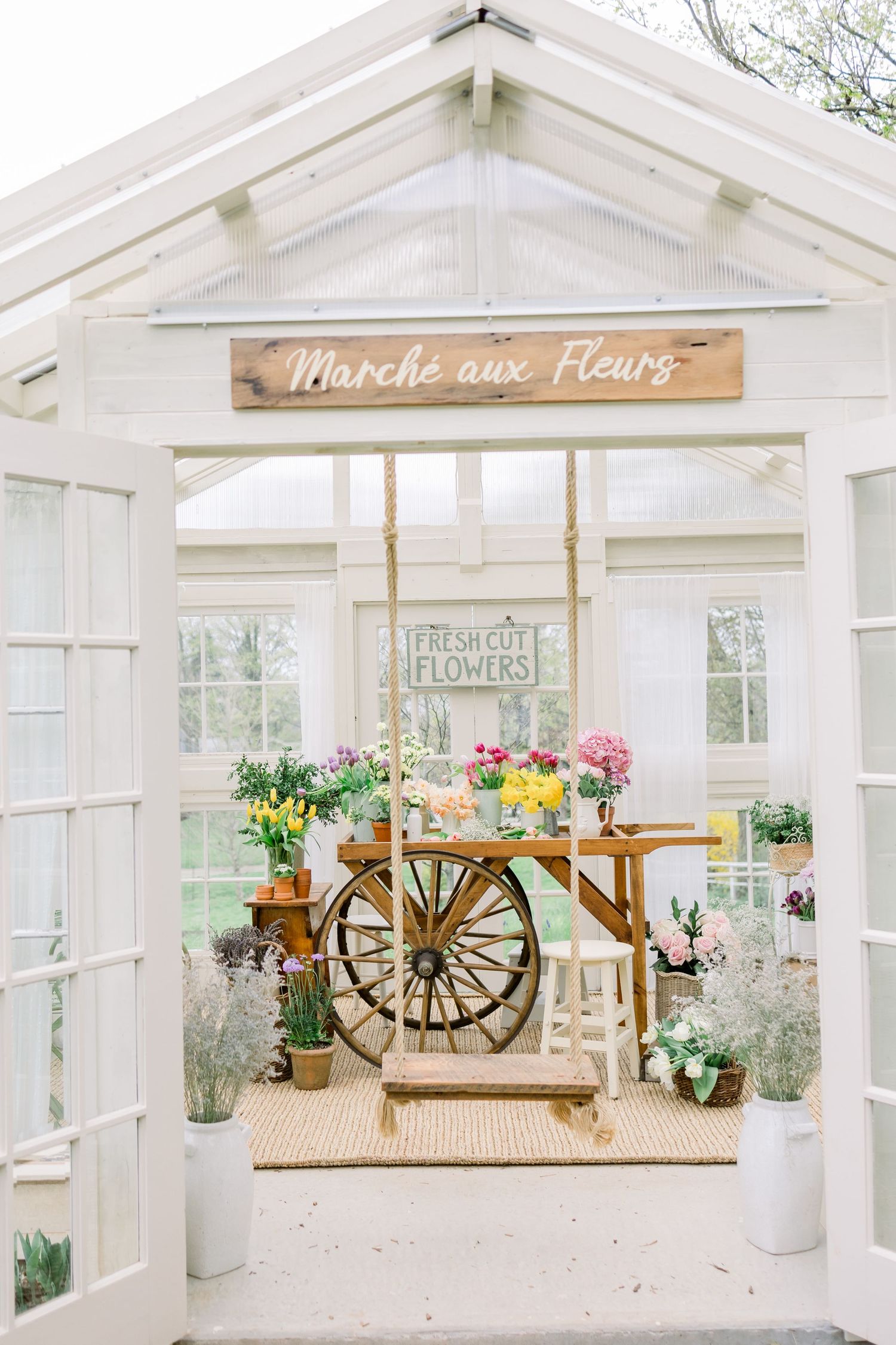 Charming French flower market cart display in a white greenhouse with 'Marche aux Fleurs' wooden sign.