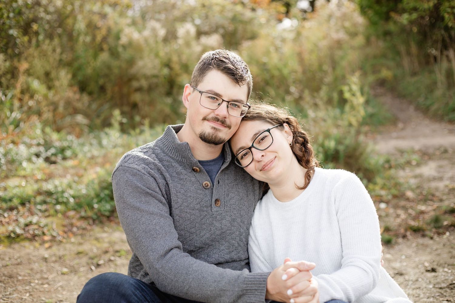 A couple wearing glasses embrace while sitting outdoors on a dirt path surrounded by autumn foliage.