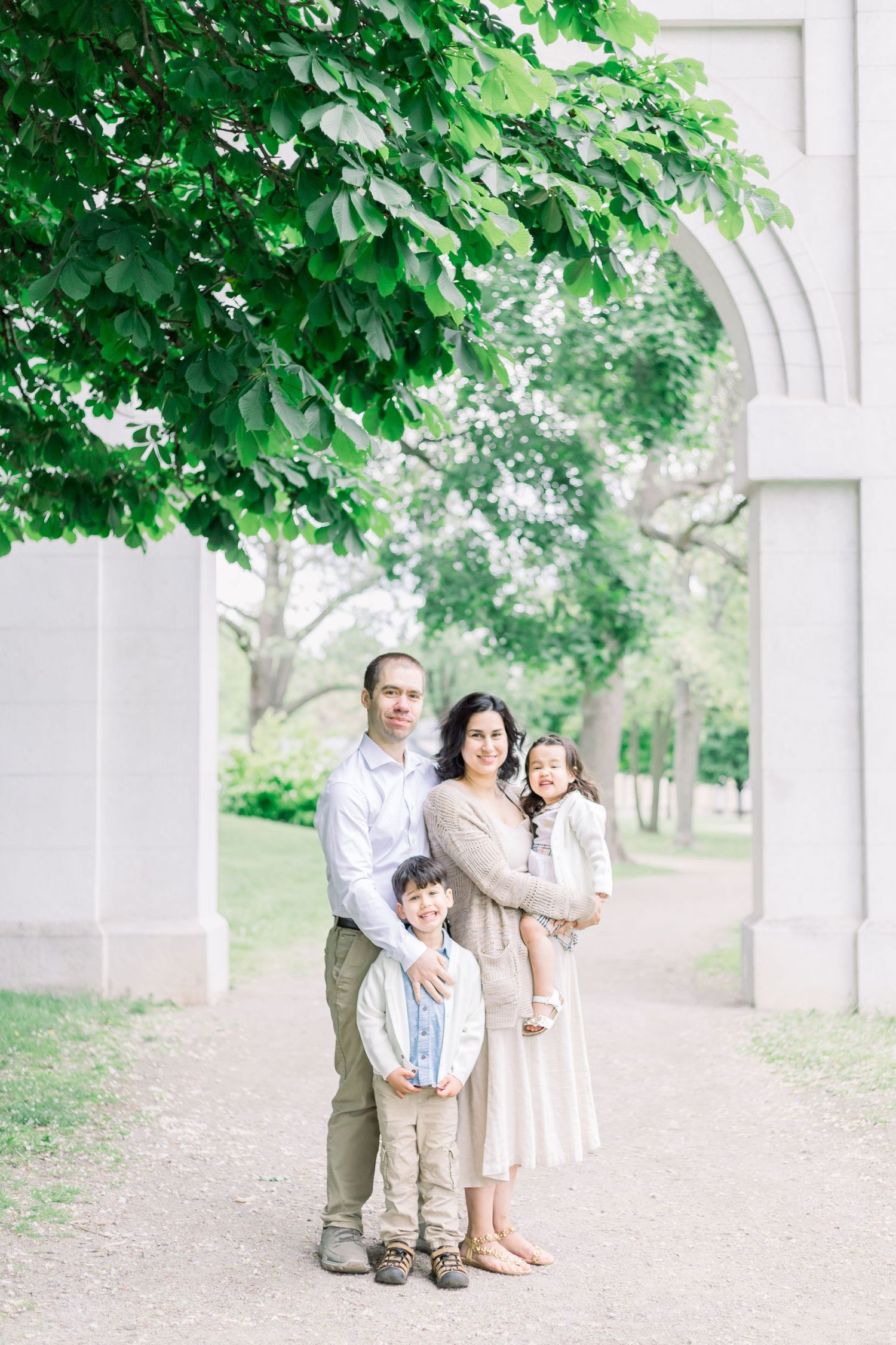 A family group poses against a white architectural feature surrounded by lush green trees in a park setting.