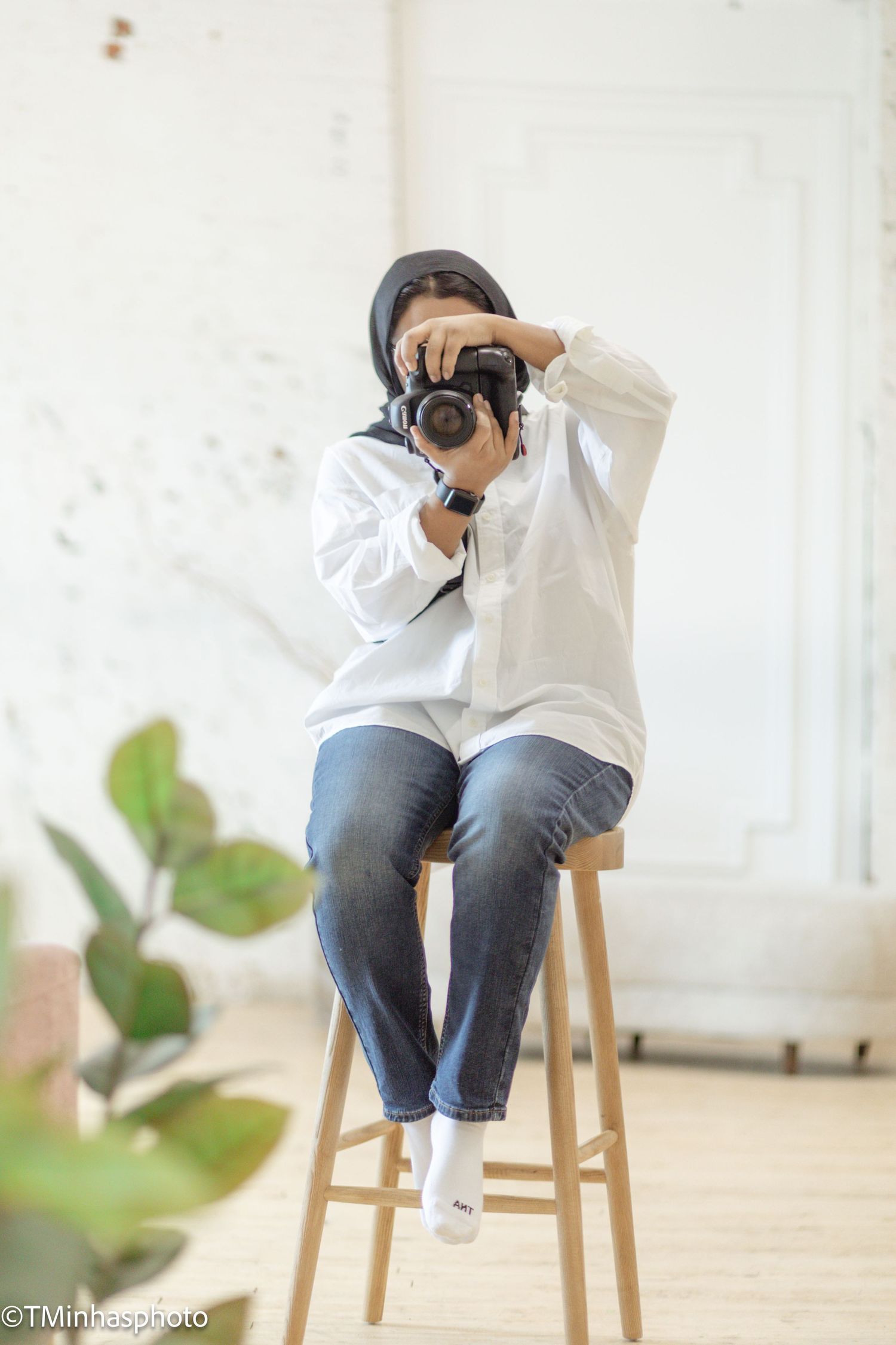 A photographer in white sweater and jeans sits on a wooden stool while taking pictures in a bright indoor setting.