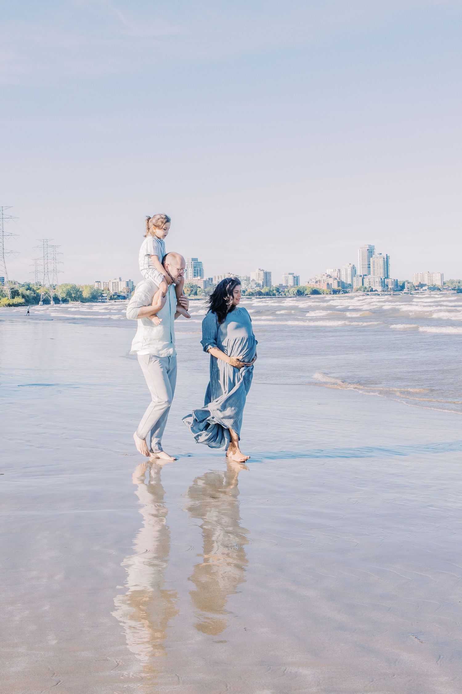 Two people enjoying a walk along a beach with city skyline in the background on a sunny day.