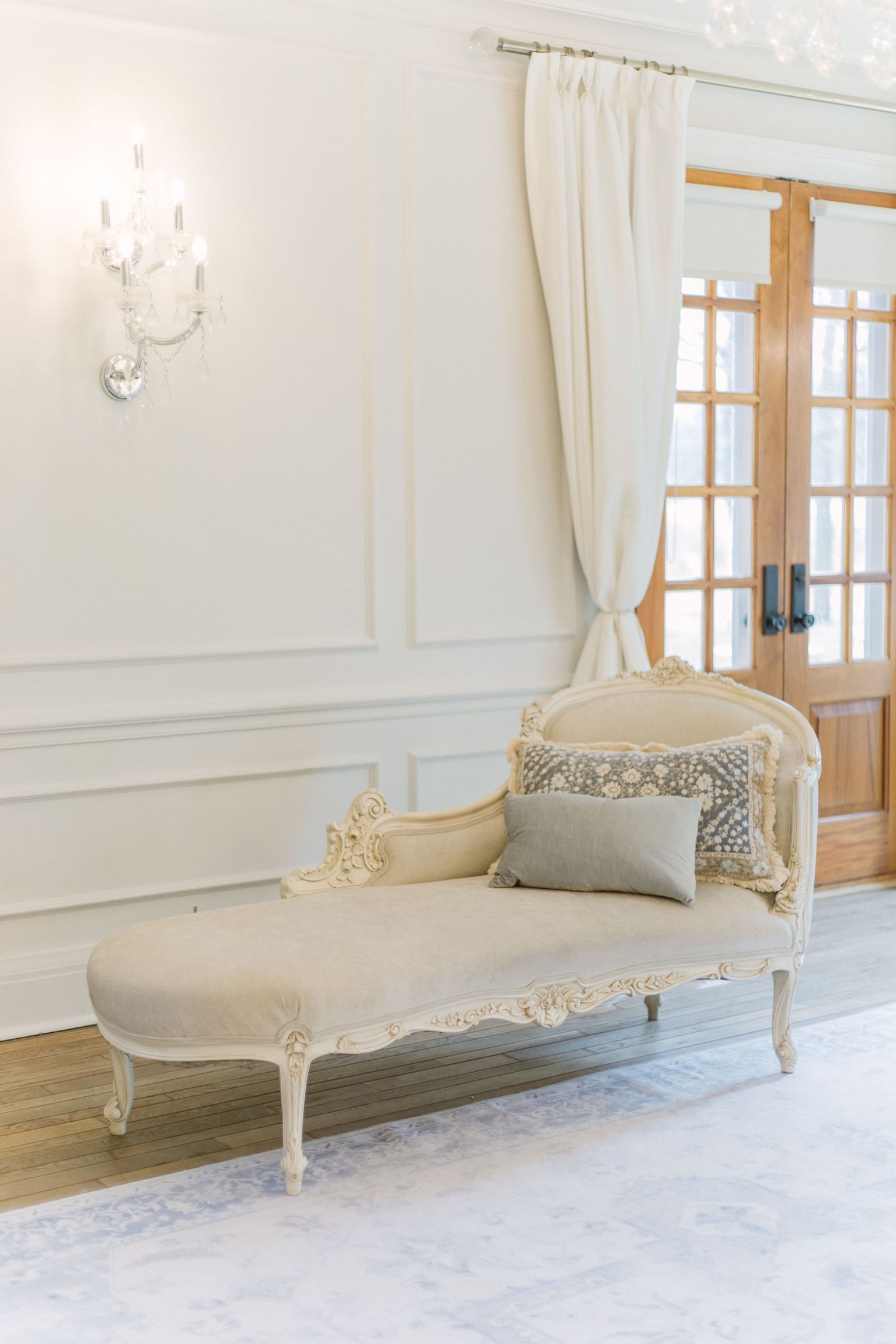 Elegant cream-colored chaise lounge with ornate carved details and gray pillow in a bright white room with French doors.