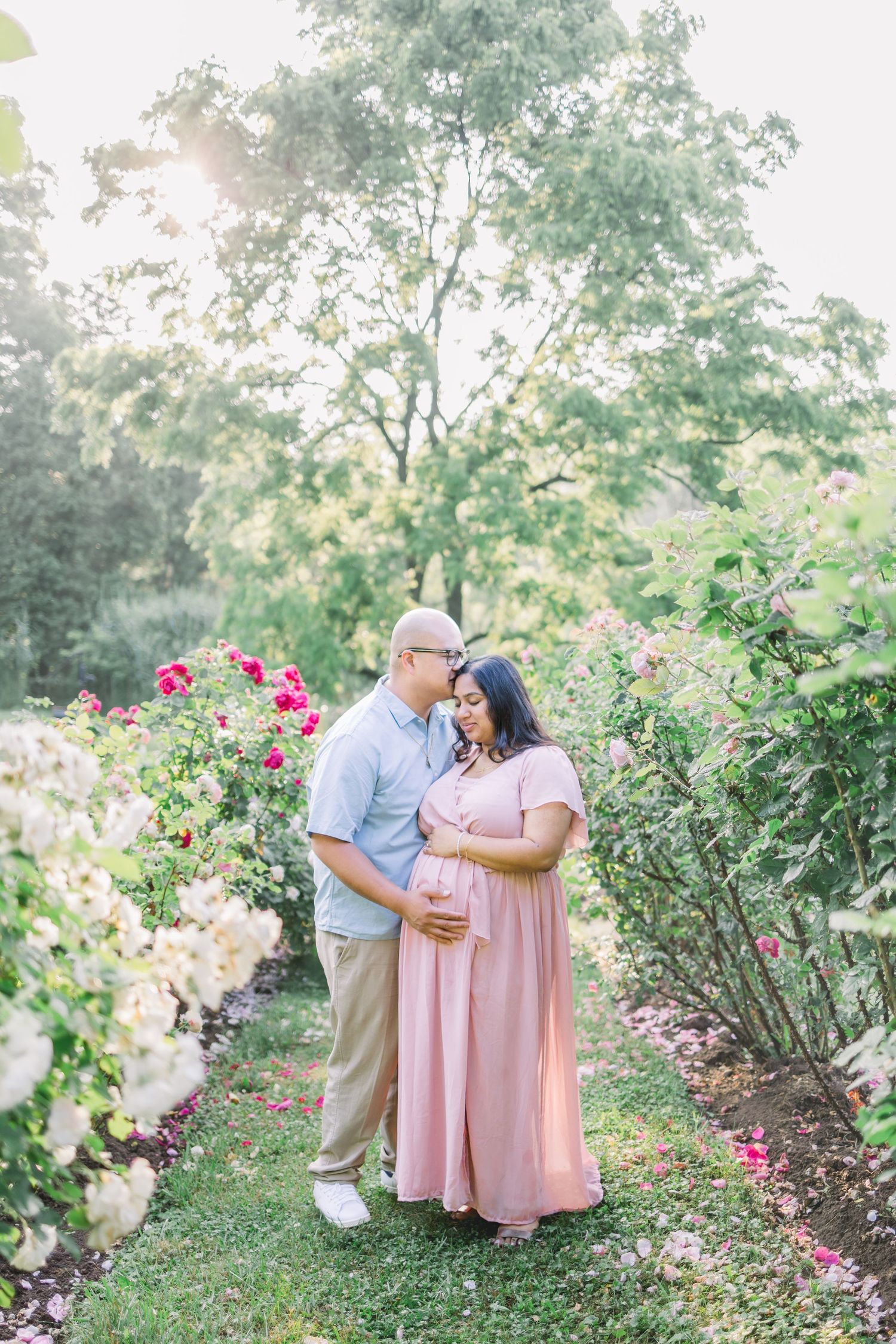 A couple shares a tender moment in a garden filled with blooming pink roses and lush greenery on a sunny day.