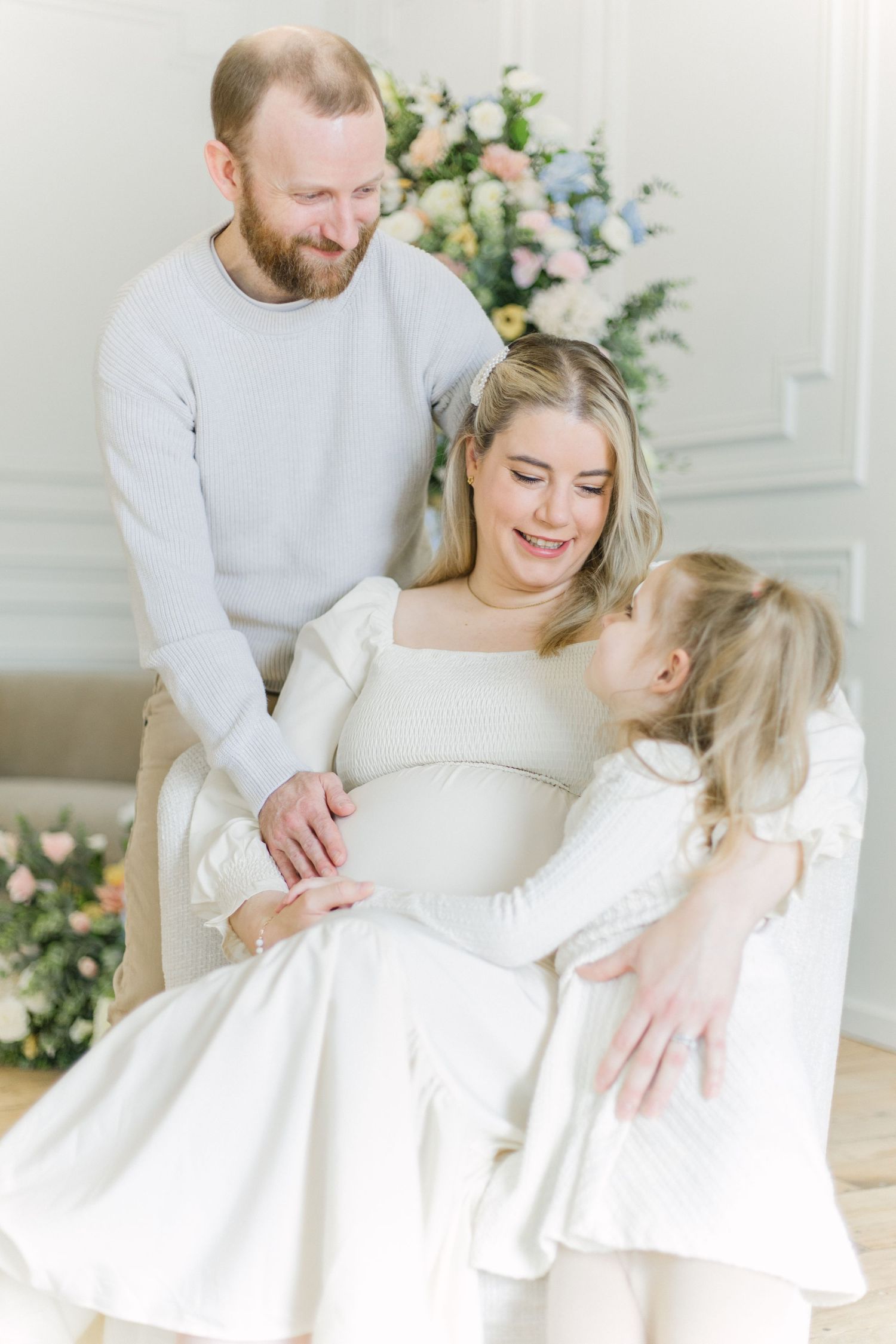 A couple poses together in matching white outfits for a romantic maternity photoshoot with floral decorations.