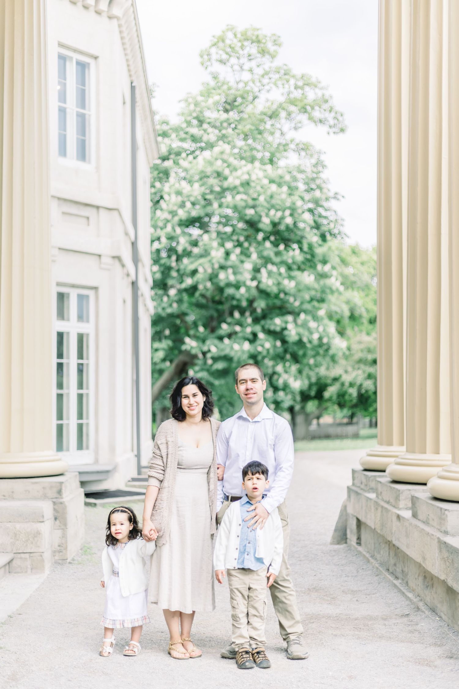 A family in white attire stands gracefully among elegant stone pillars and columns of a classical building exterior.