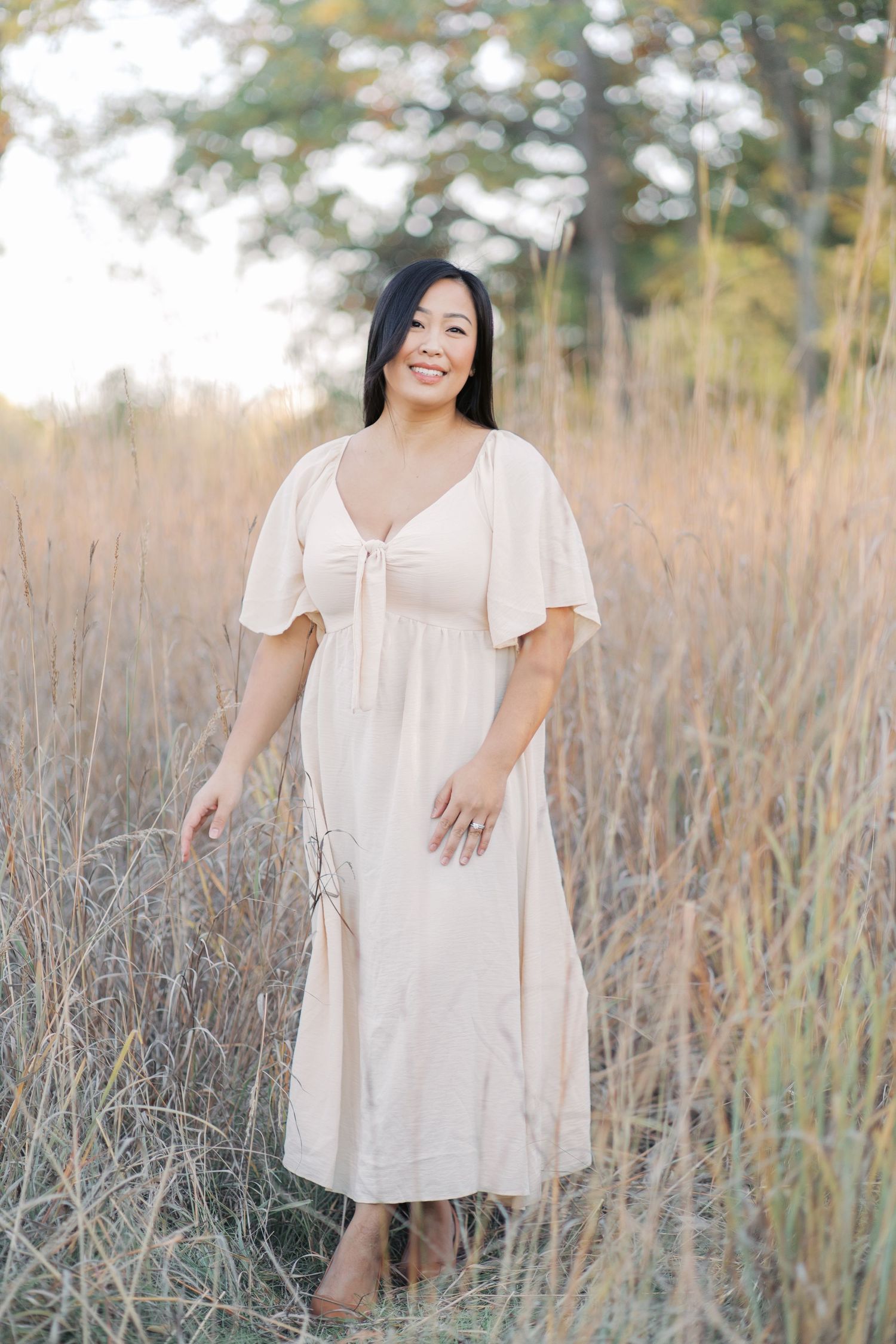 A person in a cream-colored dress poses gracefully in a field of tall grass during golden hour.
