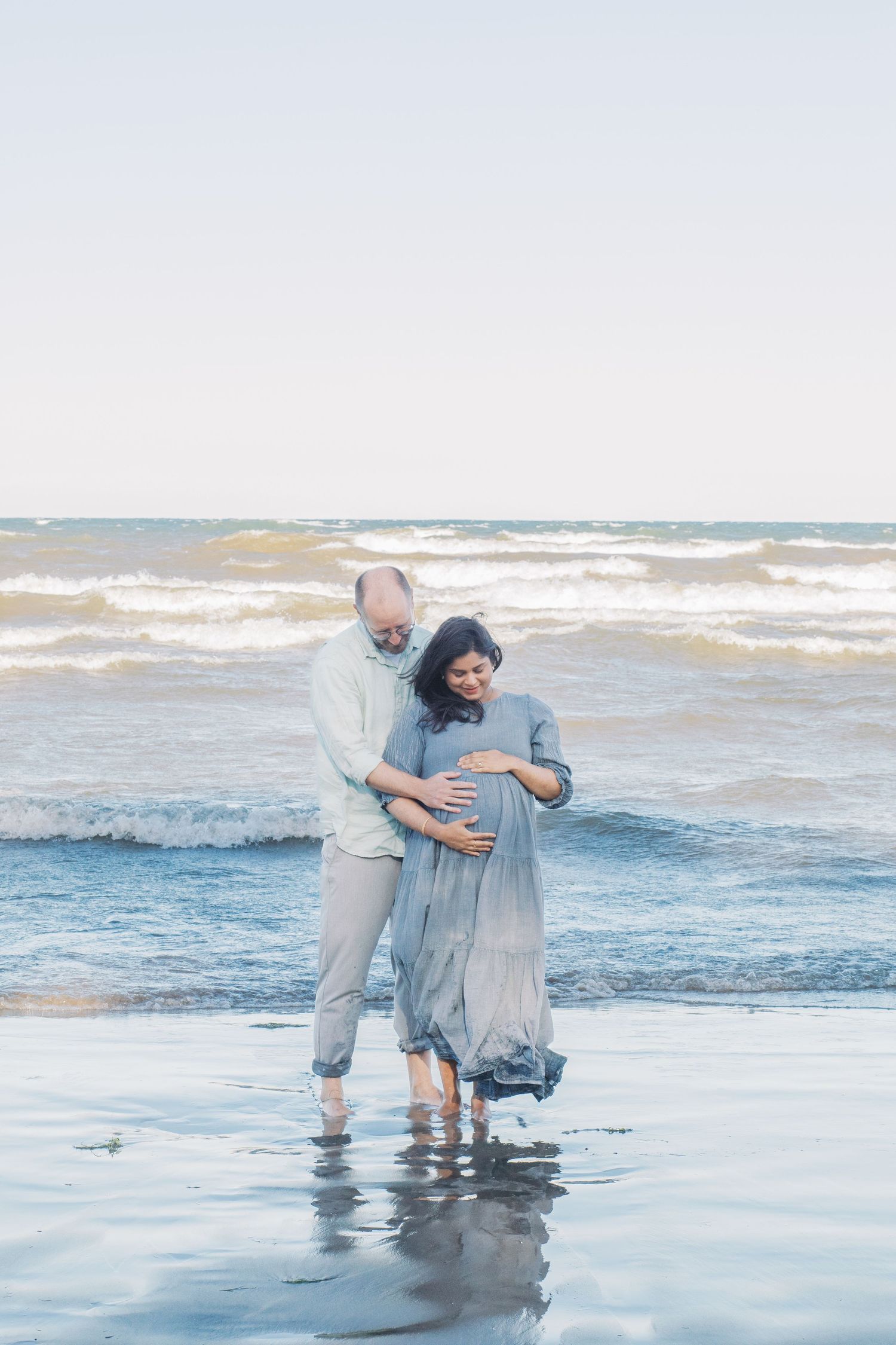 Two people embrace while standing in the shallow ocean waves wearing light colored clothing.