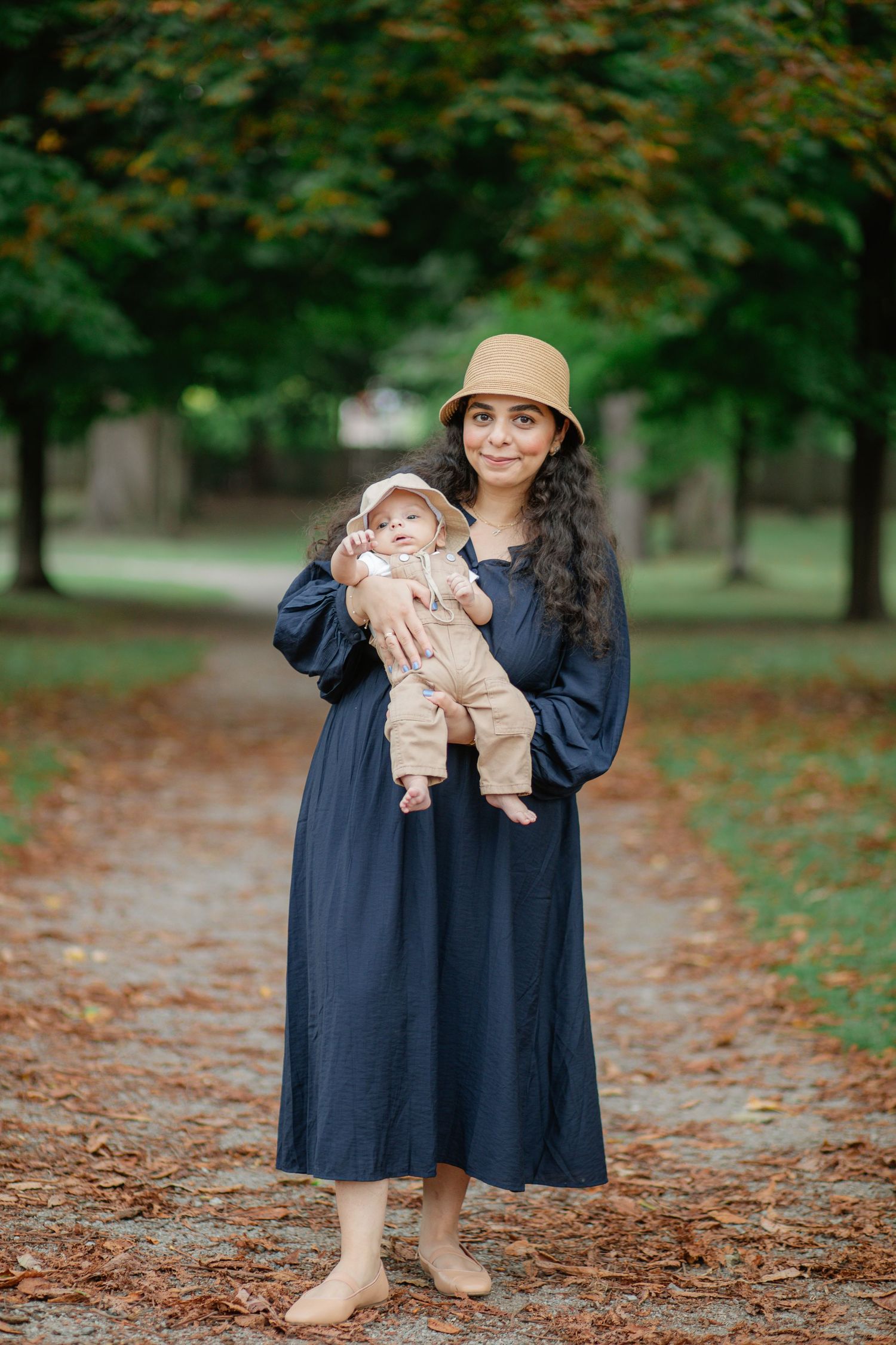 Woman in blue dress and hat holds baby while standing on fall leaf-covered path in a park.