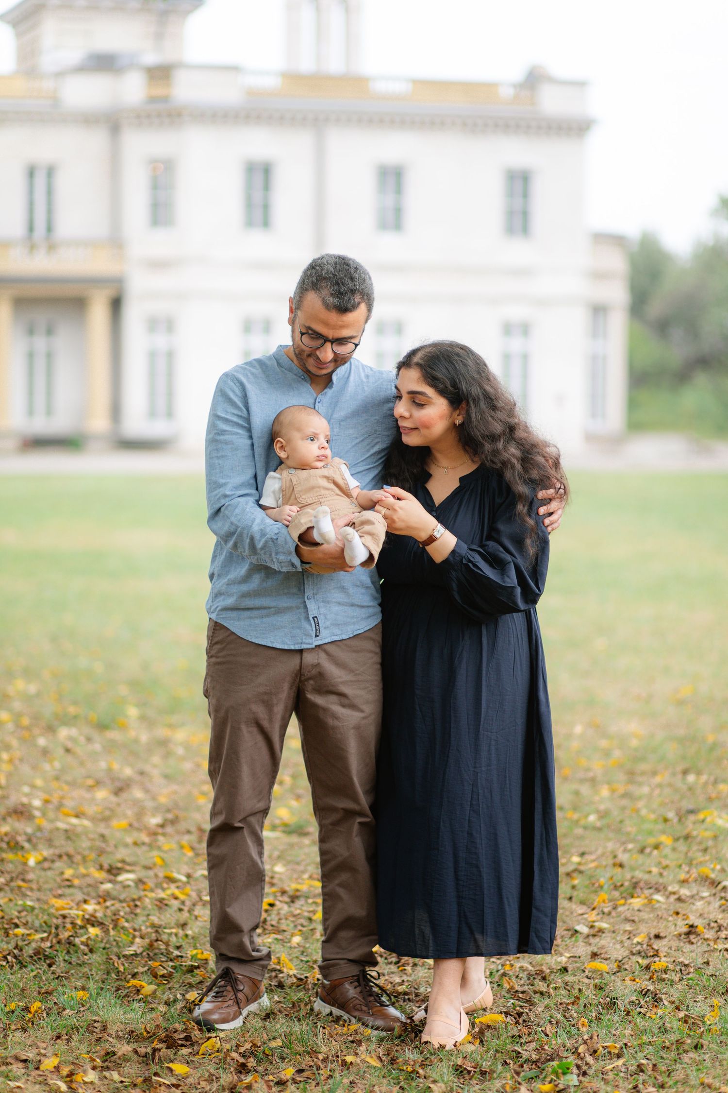 A couple stands with their baby on an autumn lawn in front of a stately white mansion.