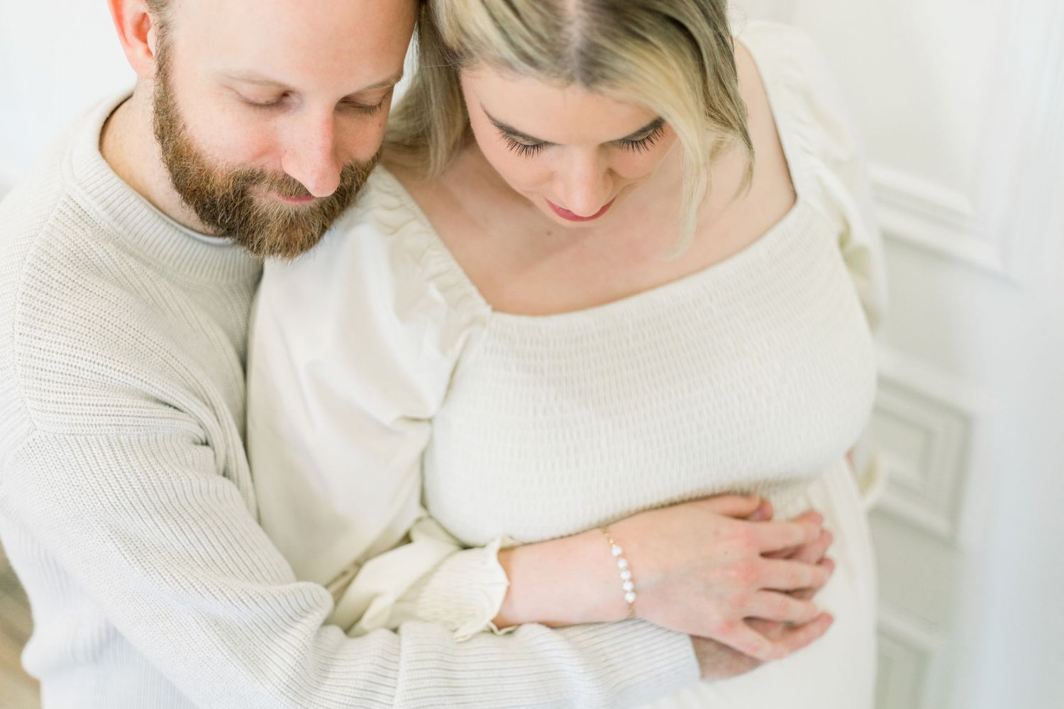A couple in soft white clothing embraces tenderly during an intimate photo session against a light background.