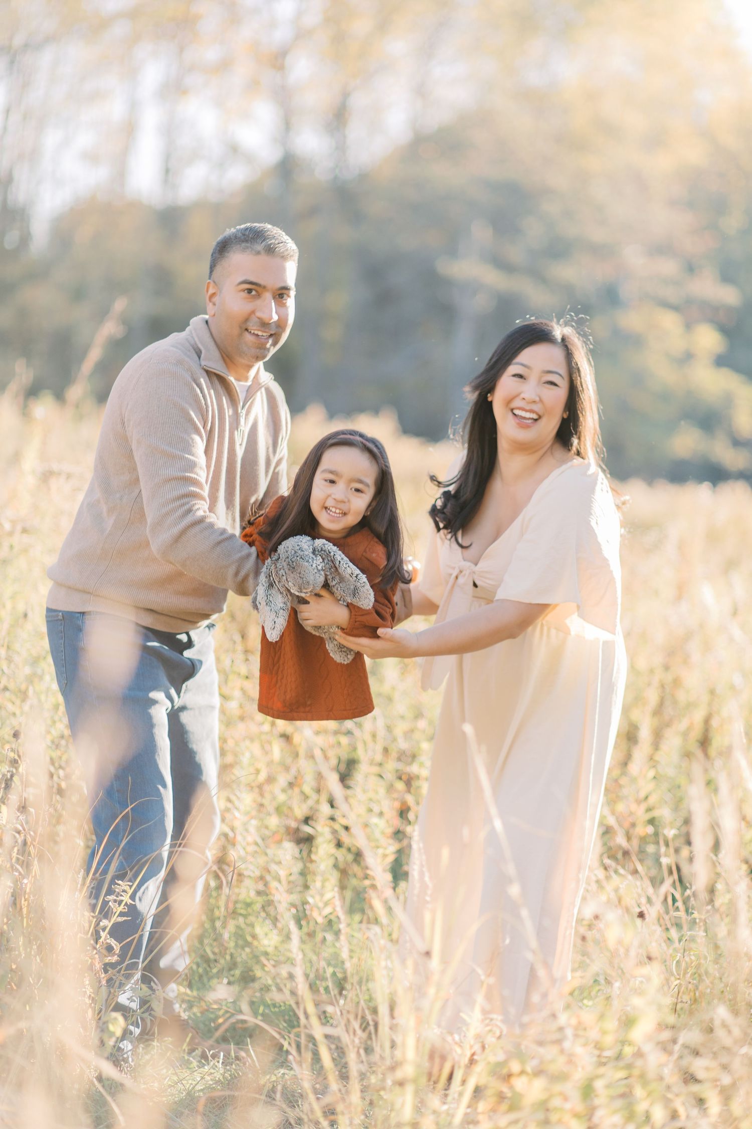A couple in casual attire plays and laughs together in a sunlit meadow with tall golden grass during sunset.