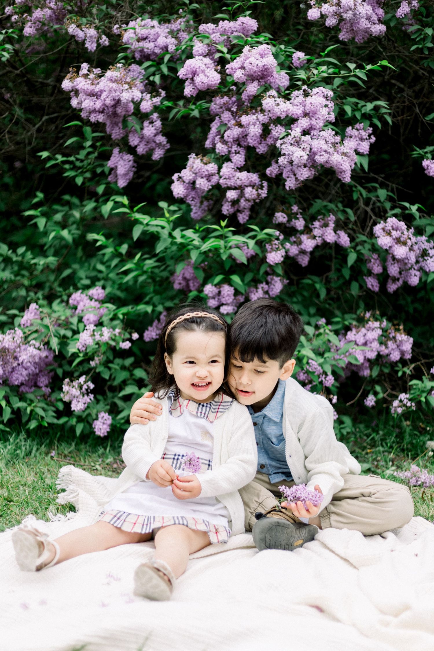 Two children sit on a blanket beneath blooming purple lilac bushes during springtime.
