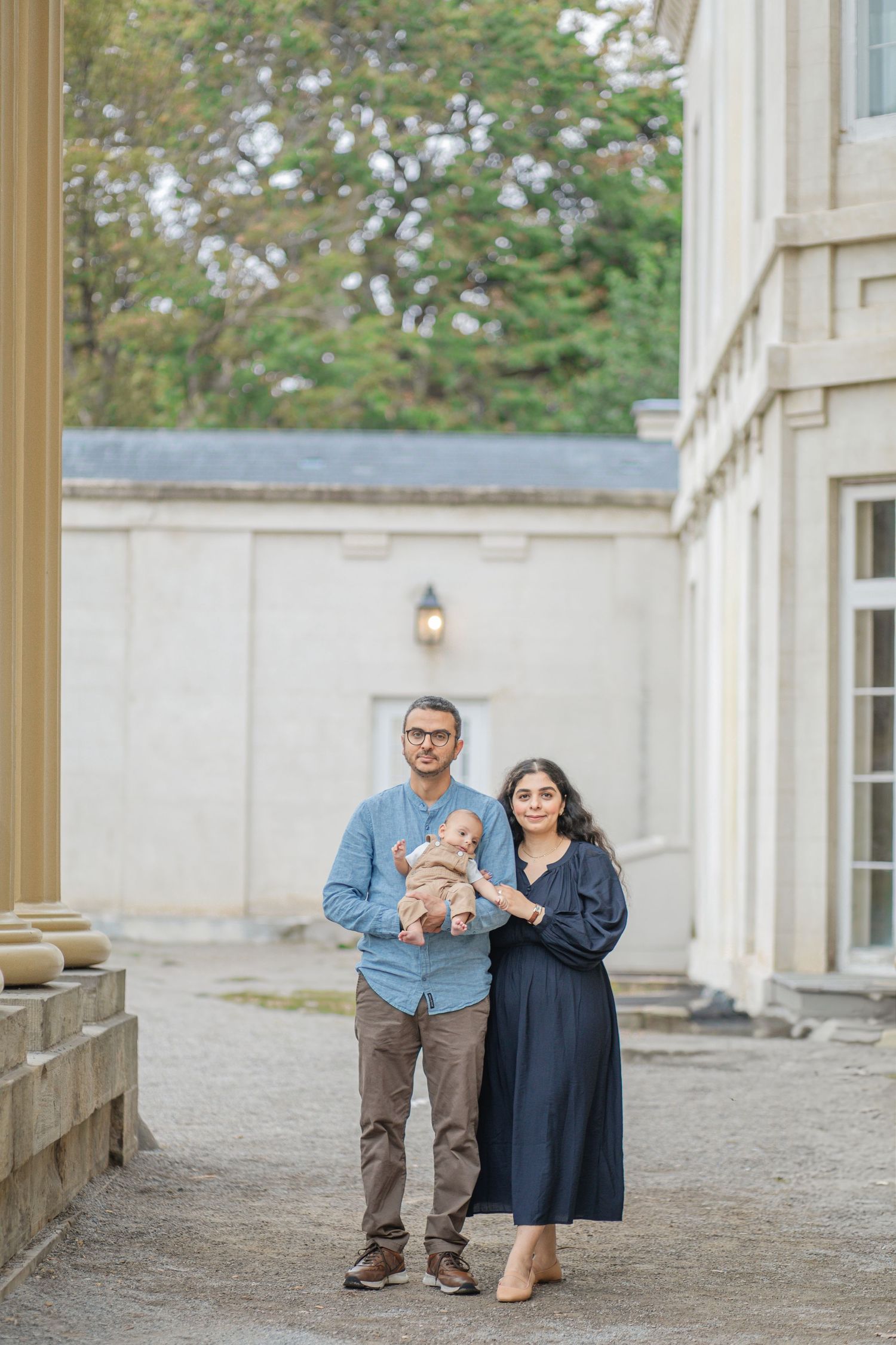 A couple poses with their baby outside of a classic stone building with a lantern on the wall.