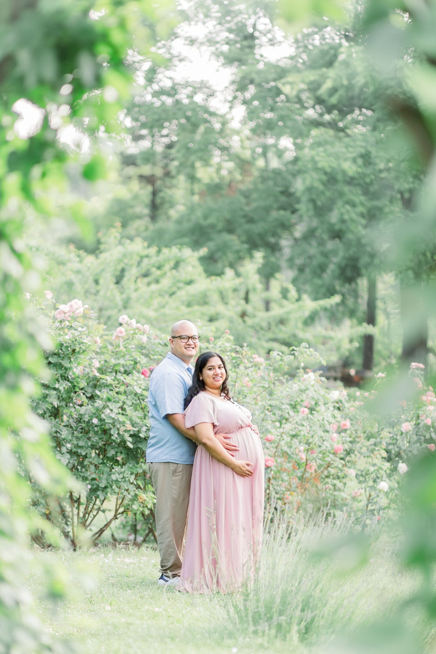 A couple stands together in a dreamy garden setting with soft green foliage and flowering bushes surrounding them.