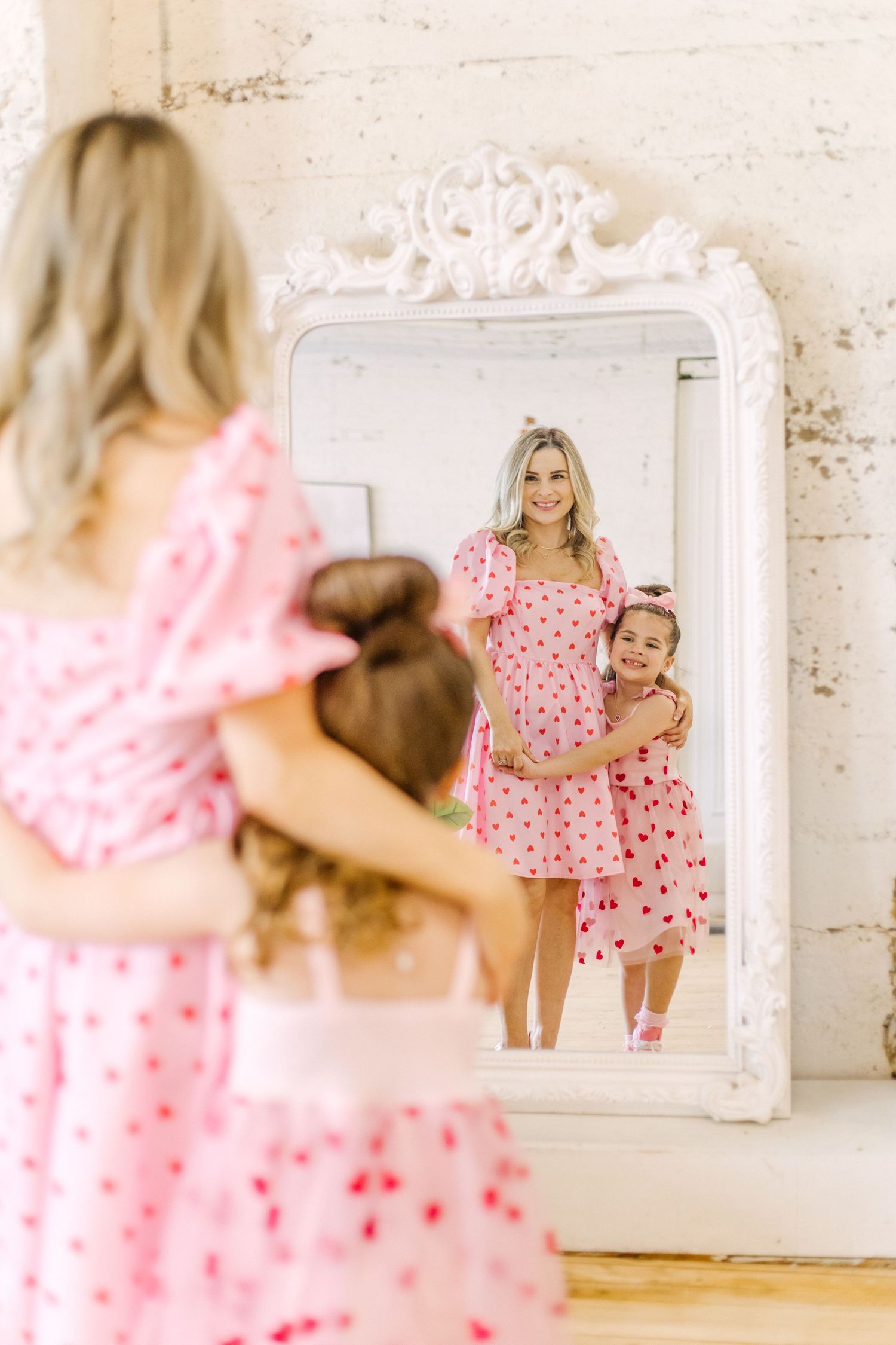 A parent and child wearing matching pink heart-patterned dresses pose together in front of an ornate white framed mirror.