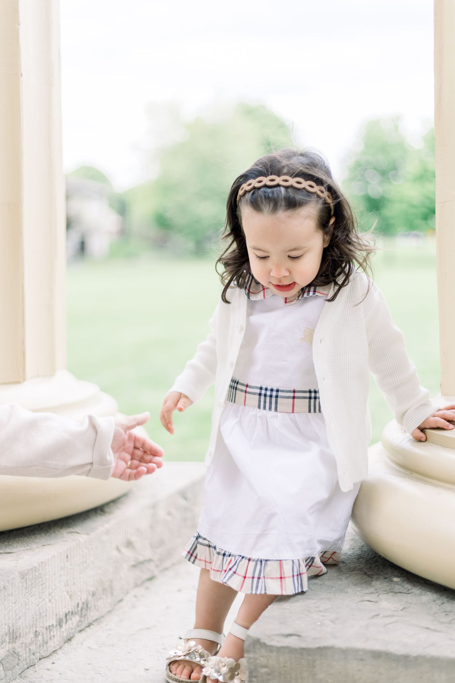 Young child wearing a white dress with plaid trim playfully dances on a stone ledge in a garden setting.