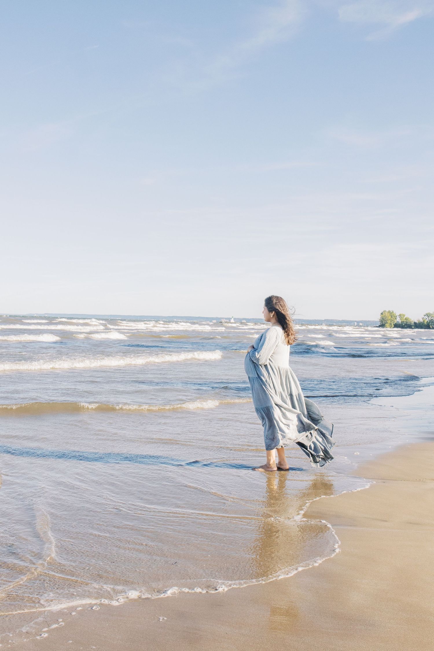Person in light clothing walks along a sandy beach shoreline as waves roll in on a bright sunny day.