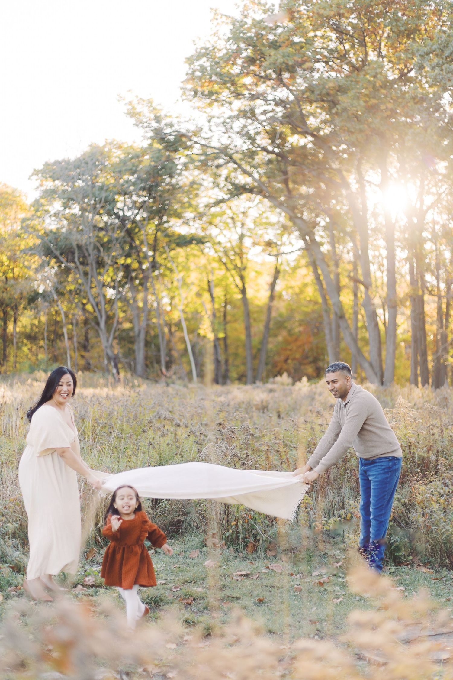 A family shares a joyful moment in a sunlit autumn forest while holding a white blanket together.