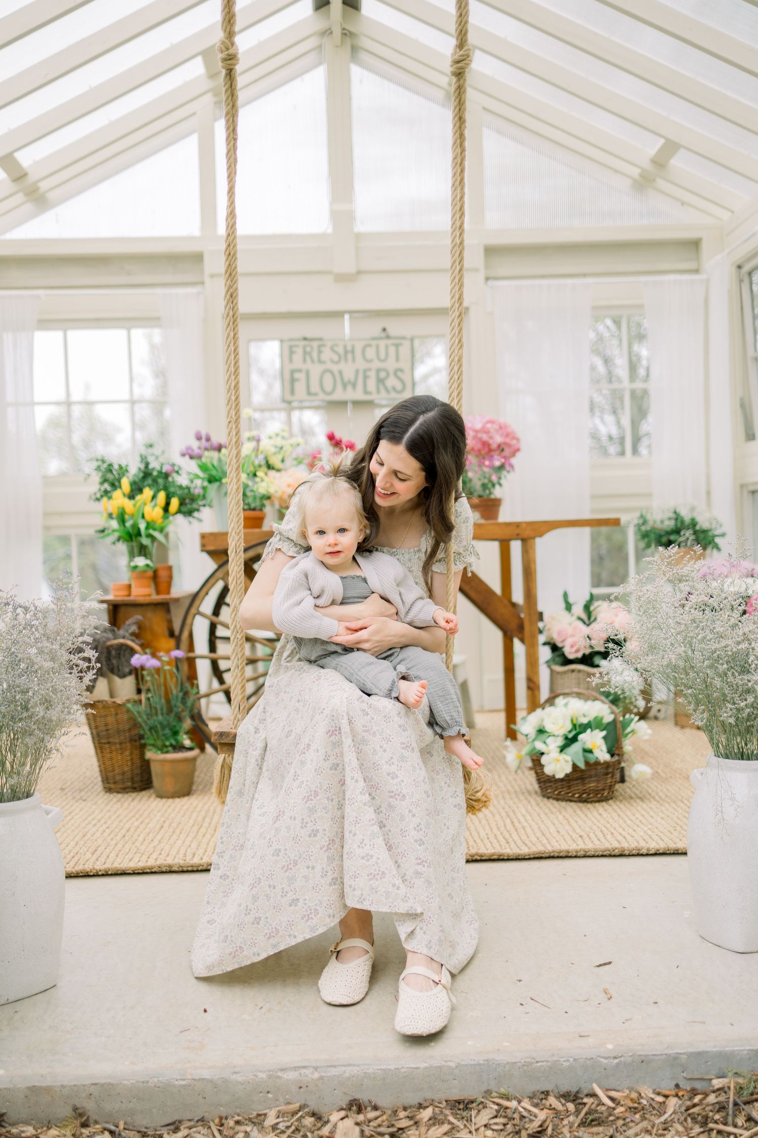 A bright sunlit greenhouse interior with white wooden furniture, potted plants, and floral decor creates a serene garden setting.