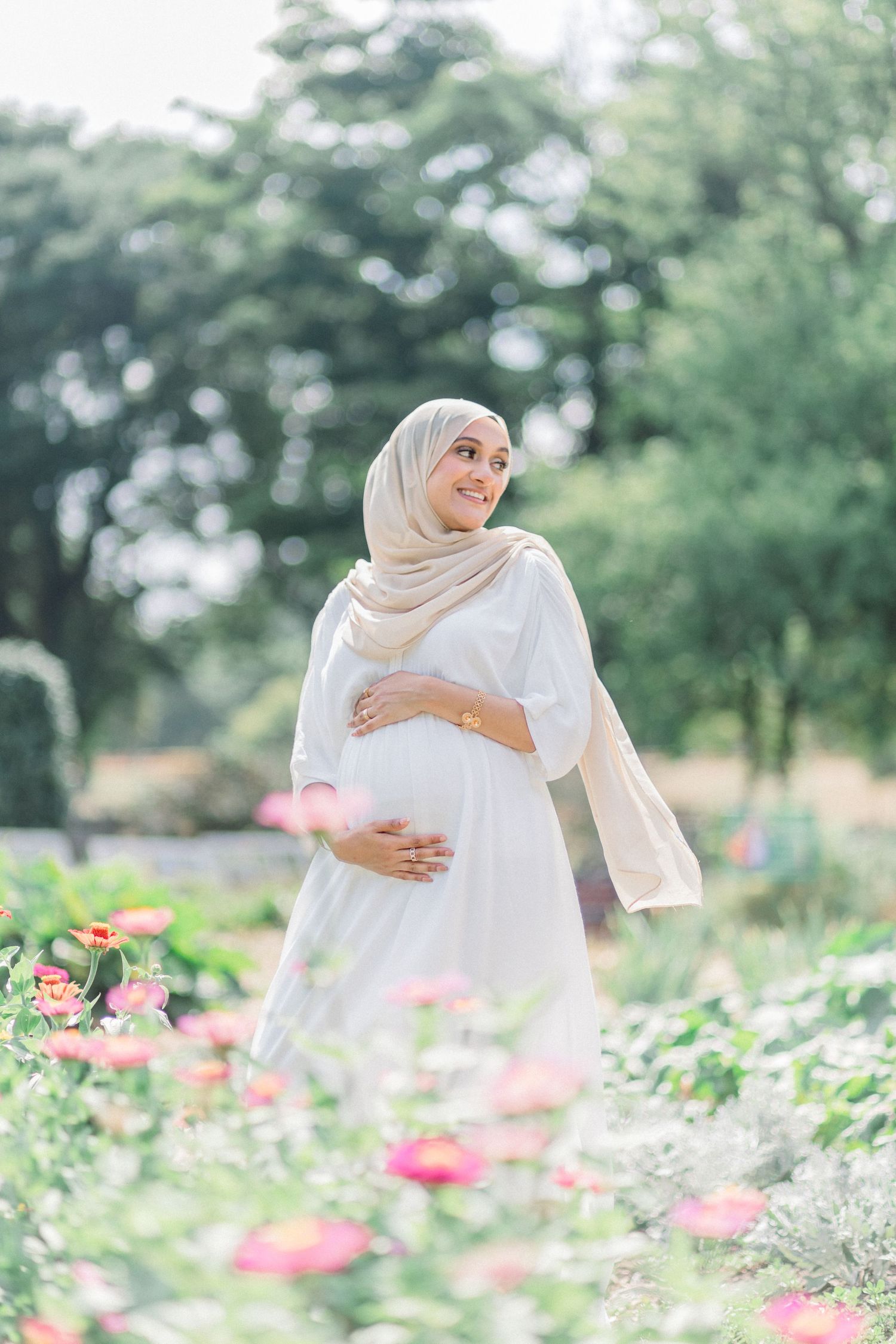 A person in a white hijab poses gracefully in a garden with blooming pink and white flowers on a sunny day.