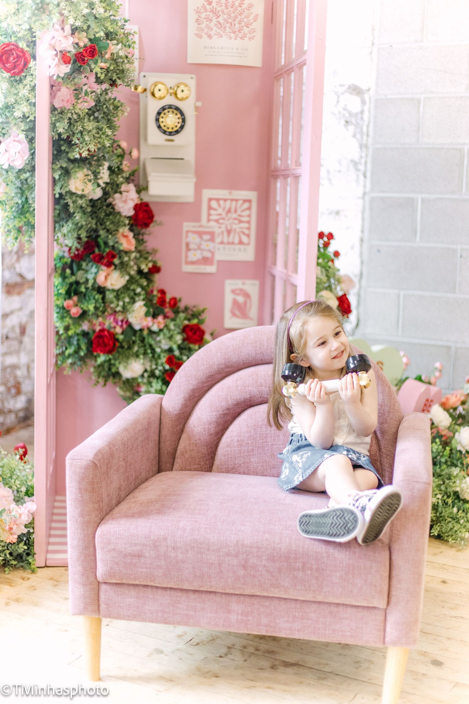 Young child sitting on pink velvet chair surrounded by floral decorations and pink walls.
