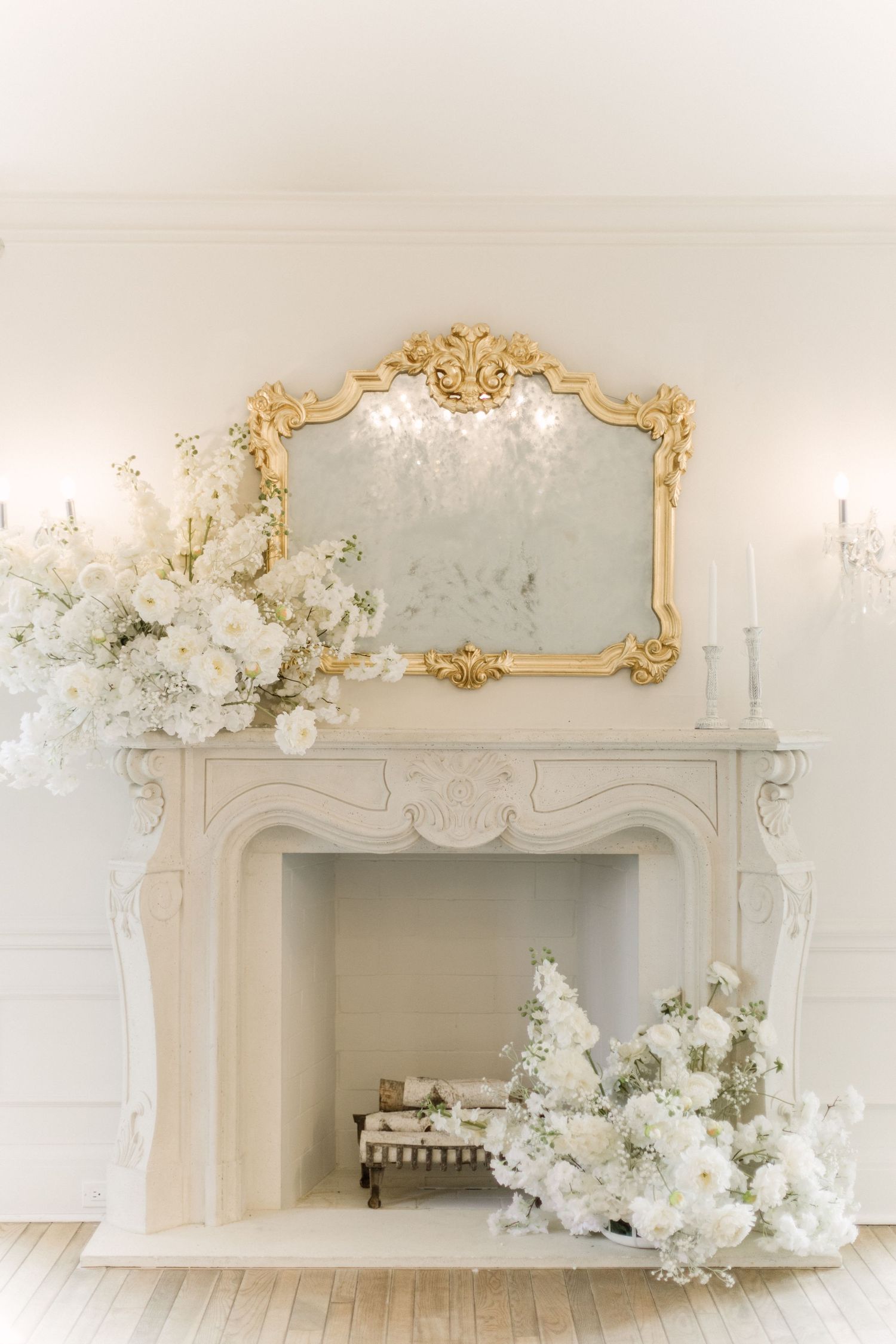 Elegant white room interior with ornate gold-framed mirror and mantle decorated with white floral arrangements.